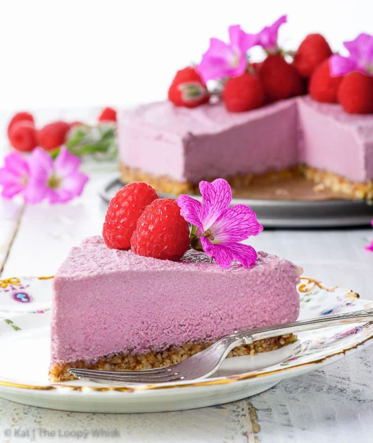 a patterened plate with a livlac colored cheesecake on top, topped with fresh raspebrries and flowers