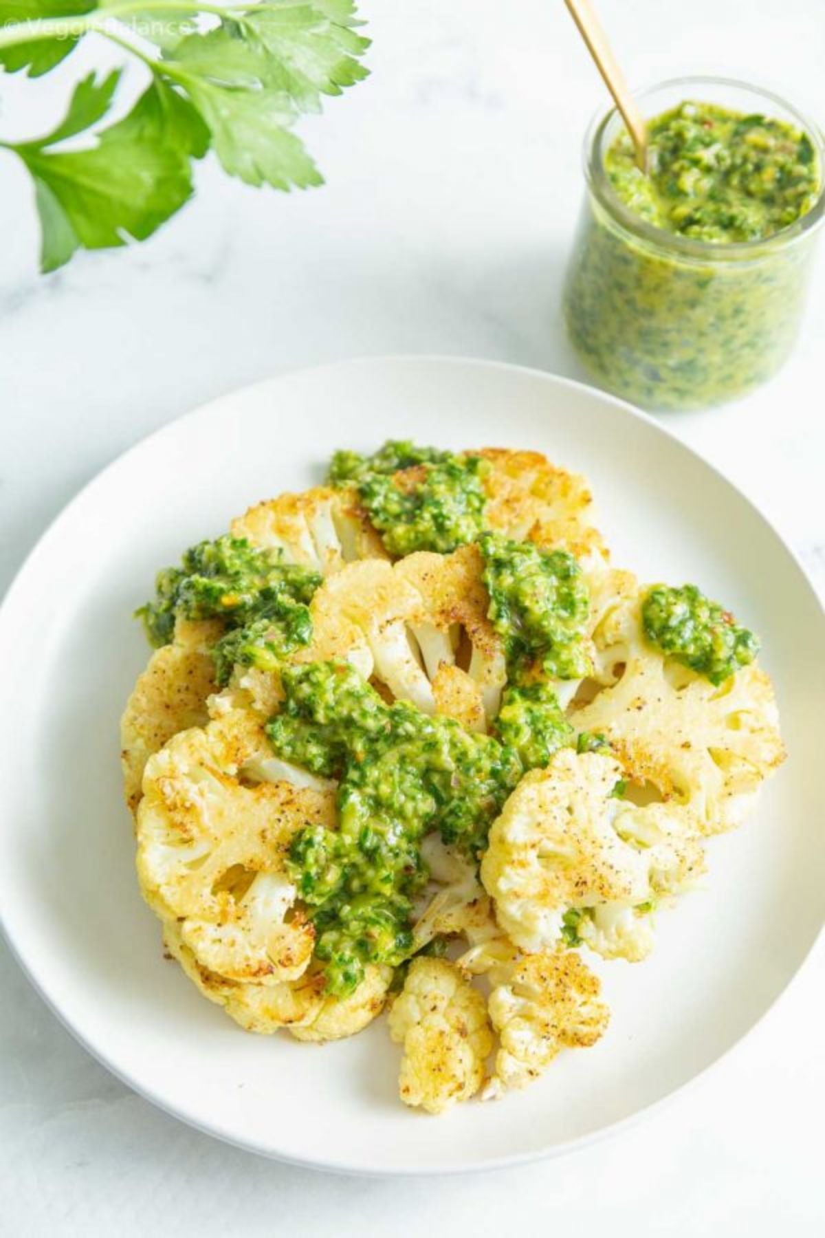 A white plate with a cauliflower steak drizzled with chimichurri. a jar of chimichurri is off to one side