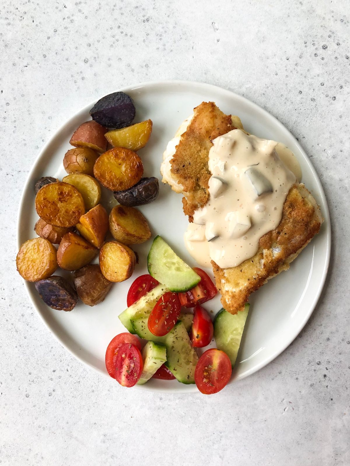 a white plate with a battered fish fillet, topped with a white sauce, roasted potatoes and a cucumber and tomato salad