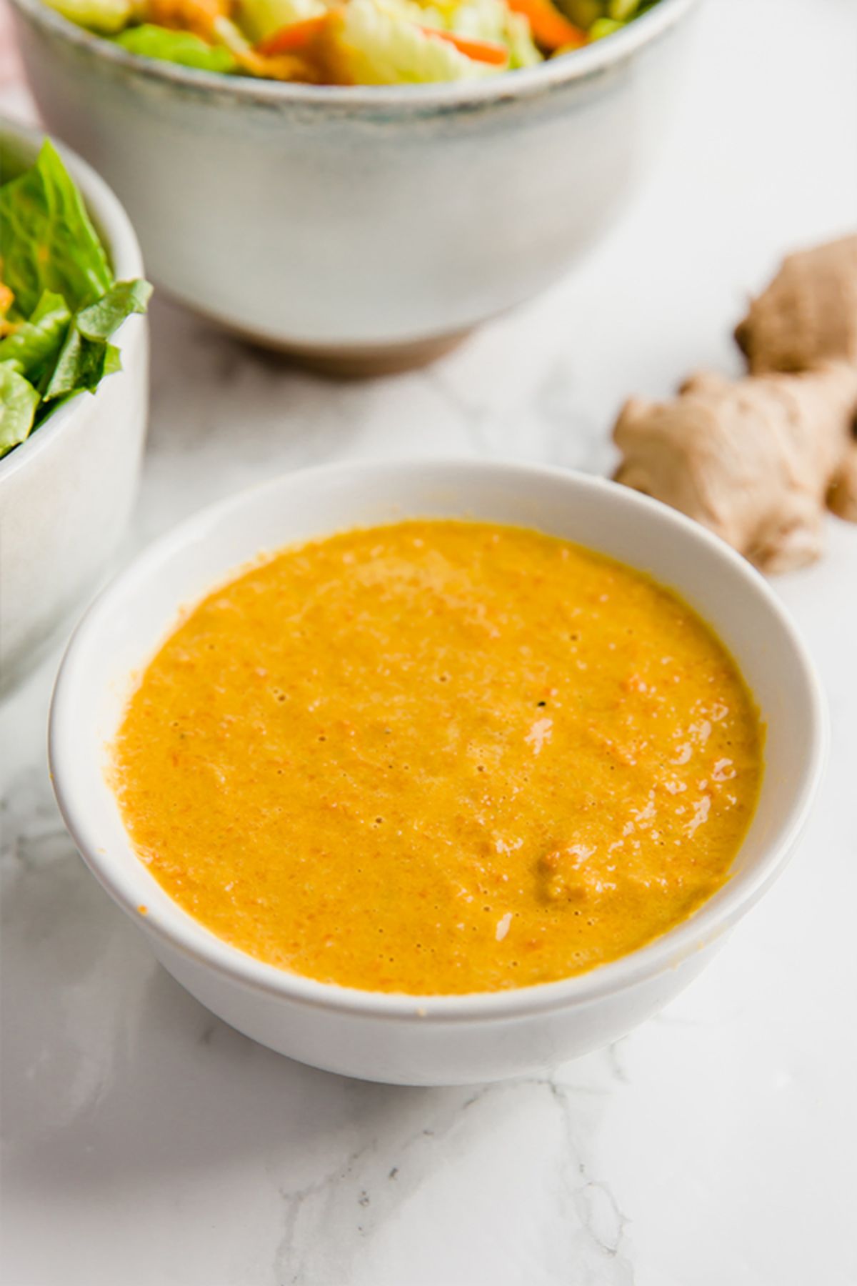 A white bowl of orange dressing next to bowls of salad and a knob of ginger