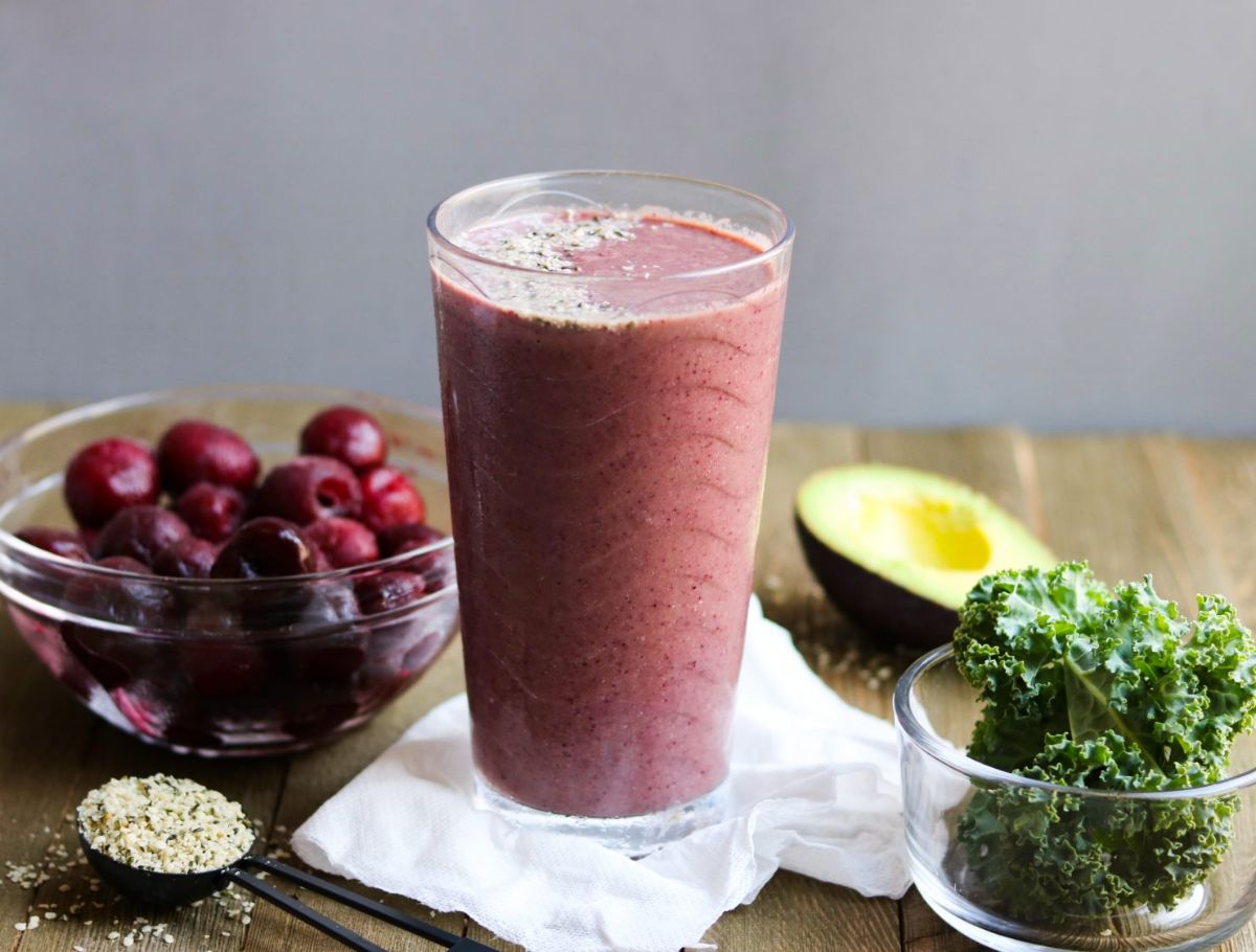 A glass of kale cherry smoothie surorunded by a bowl of cherries, a bowl of kale leaves, a spoon of hemp seeds and half an avocado