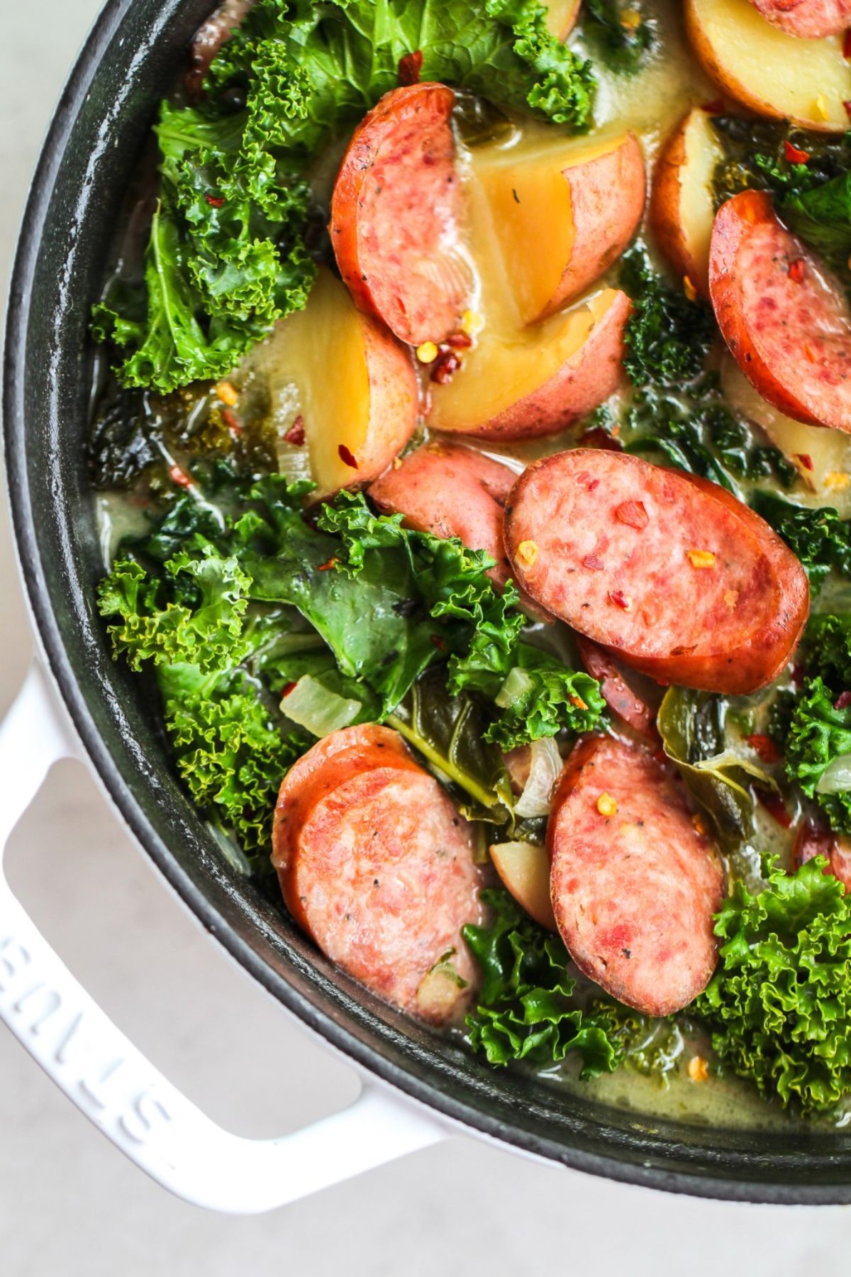 A partial shot of a cast iron casserole dish containing sausage, potato and kale soup