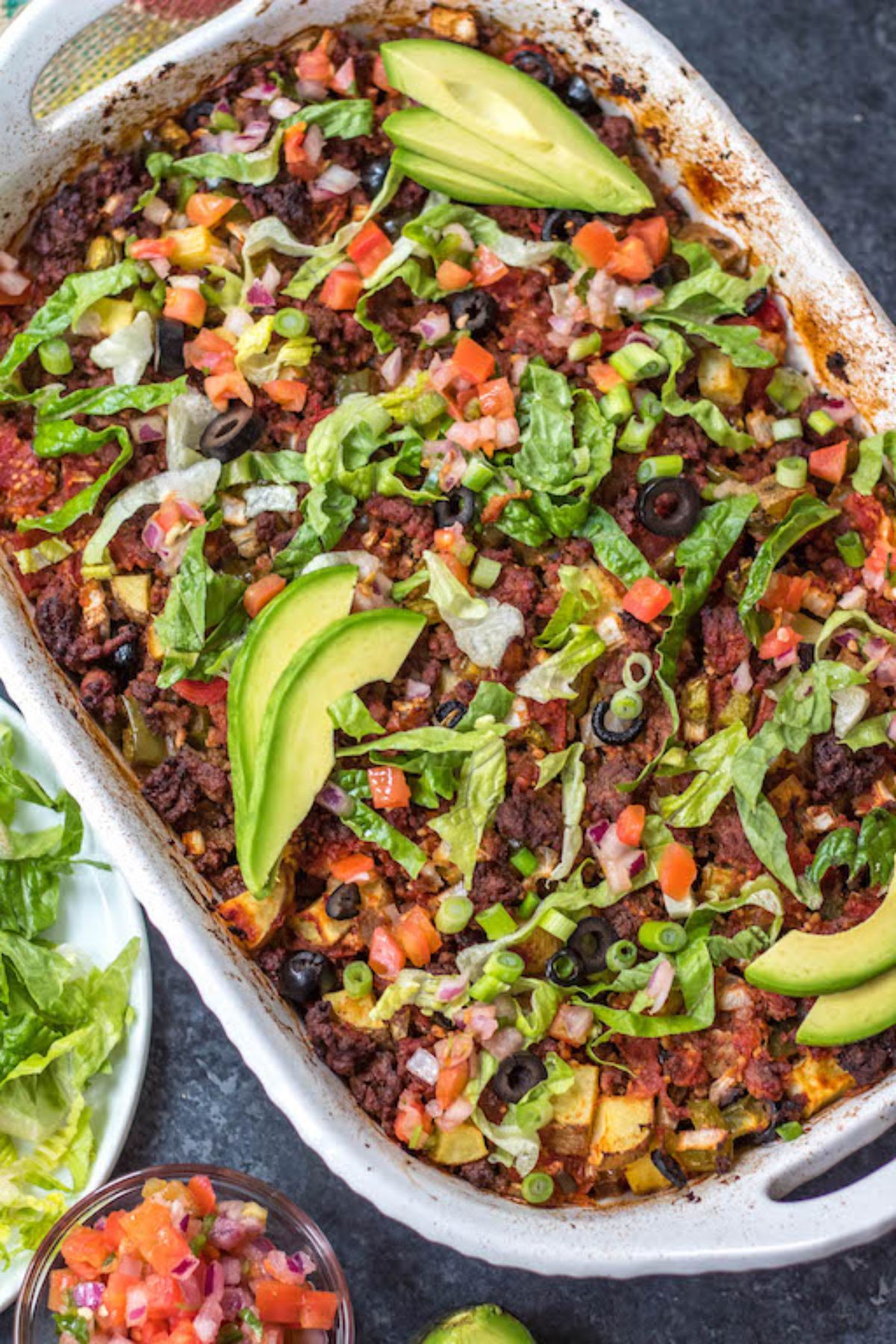 An oven dish full of beef casserole topped with avocado, lettuce, scalions and olives