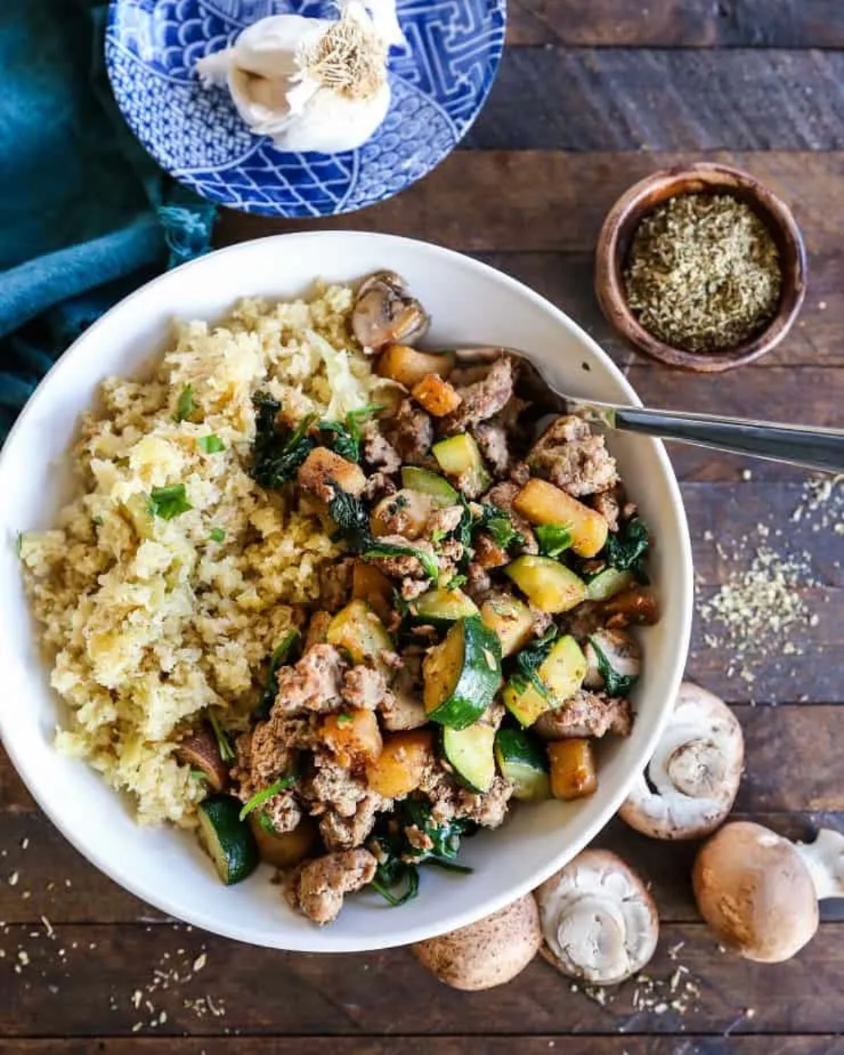 A white bowl with cauliflower rice, and a ground turkey and vegetable mixture. mushrooms, a garlic bulb and a pot of spices surround it