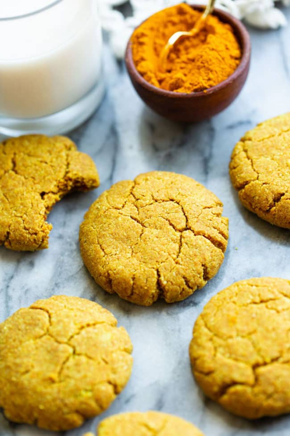 5 golden almond biscuits, one with a bit taken out of it. Behind is a glass of milk and a small pot of spice