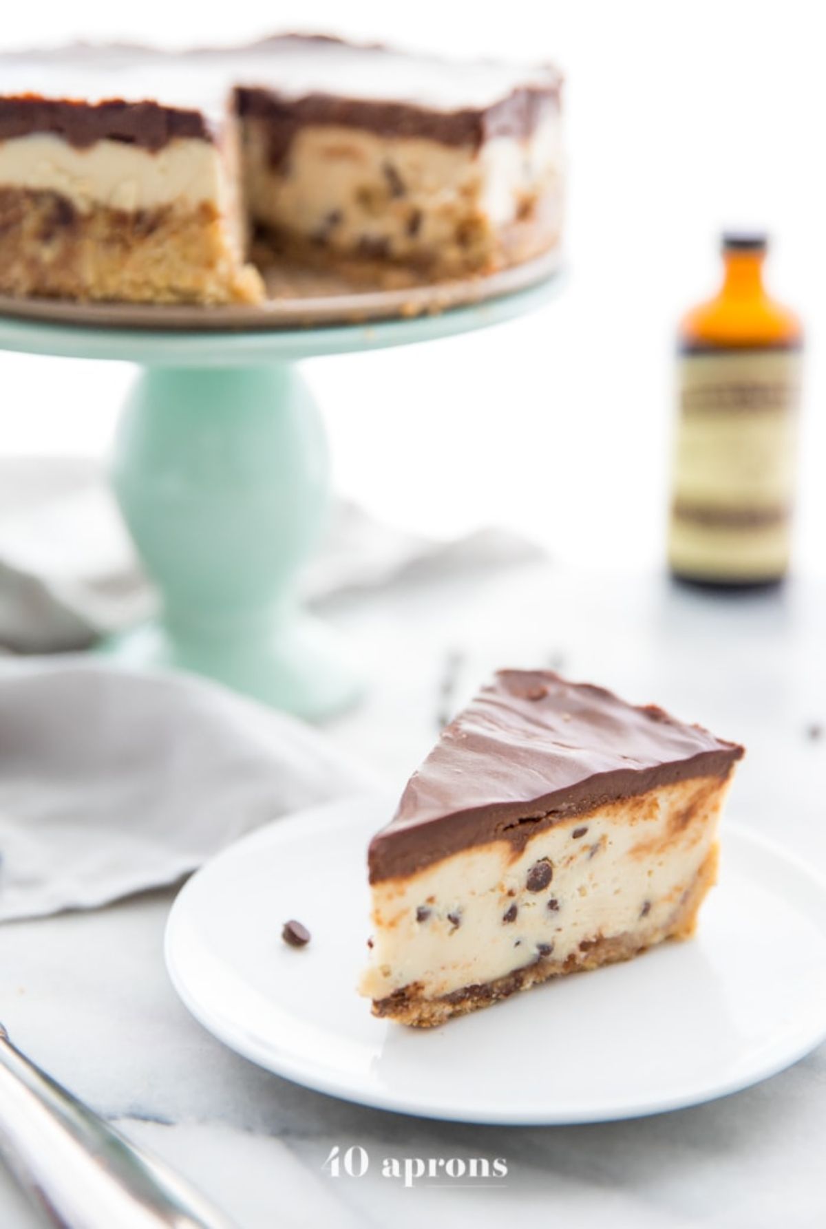 A cake stand with a layered cookie dough cheesecake with a slice cut out of it. A white palte with that slice is in front