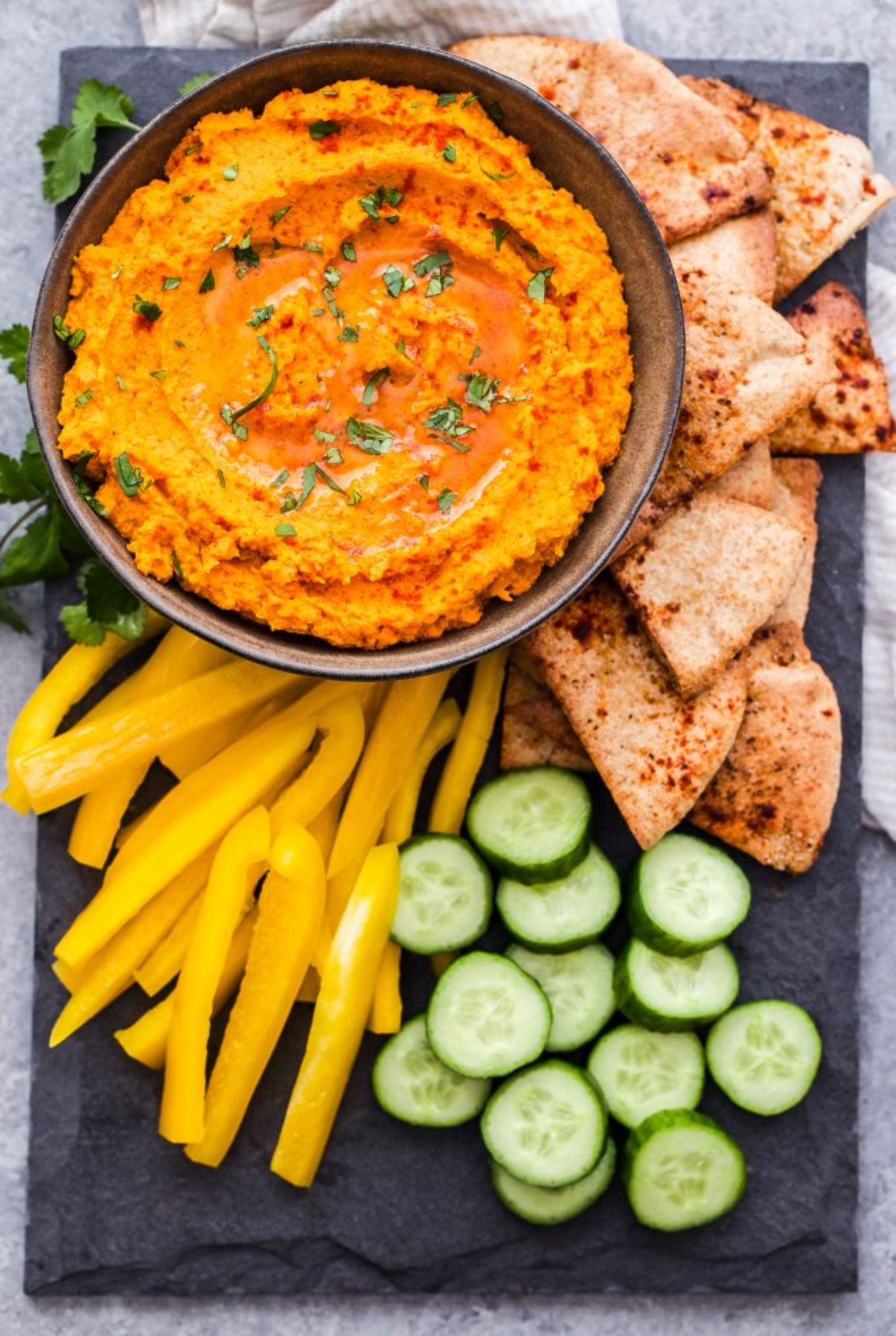 a slate tile with sliced yellow peppers, cucumbers, pitta and herbs next to a bowl of carrot and sweet potato humus