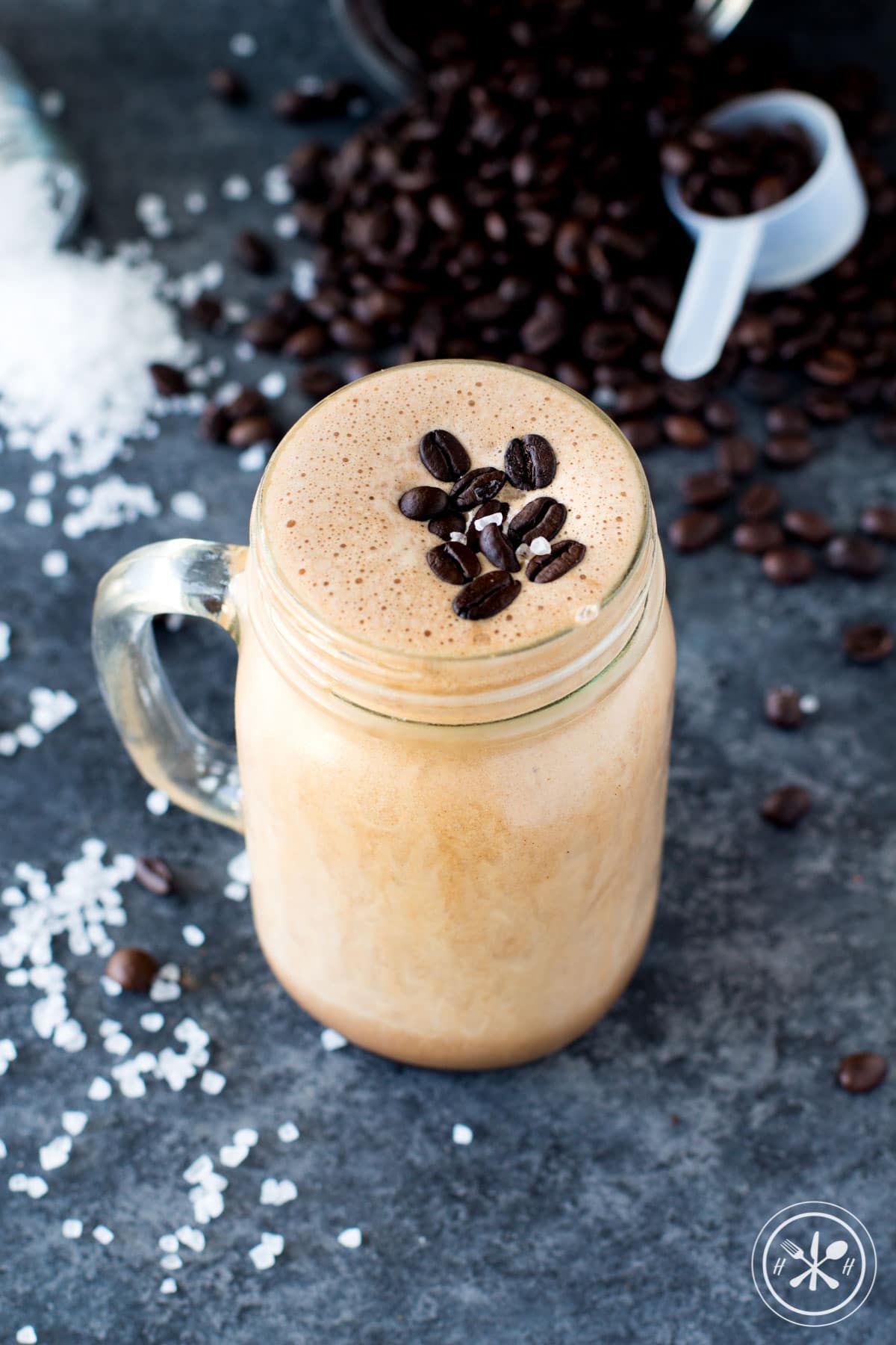a mason jar with salted mocha smoothie in it, topped with coffee beans, a pile of coffee beans and a scoop are just behind