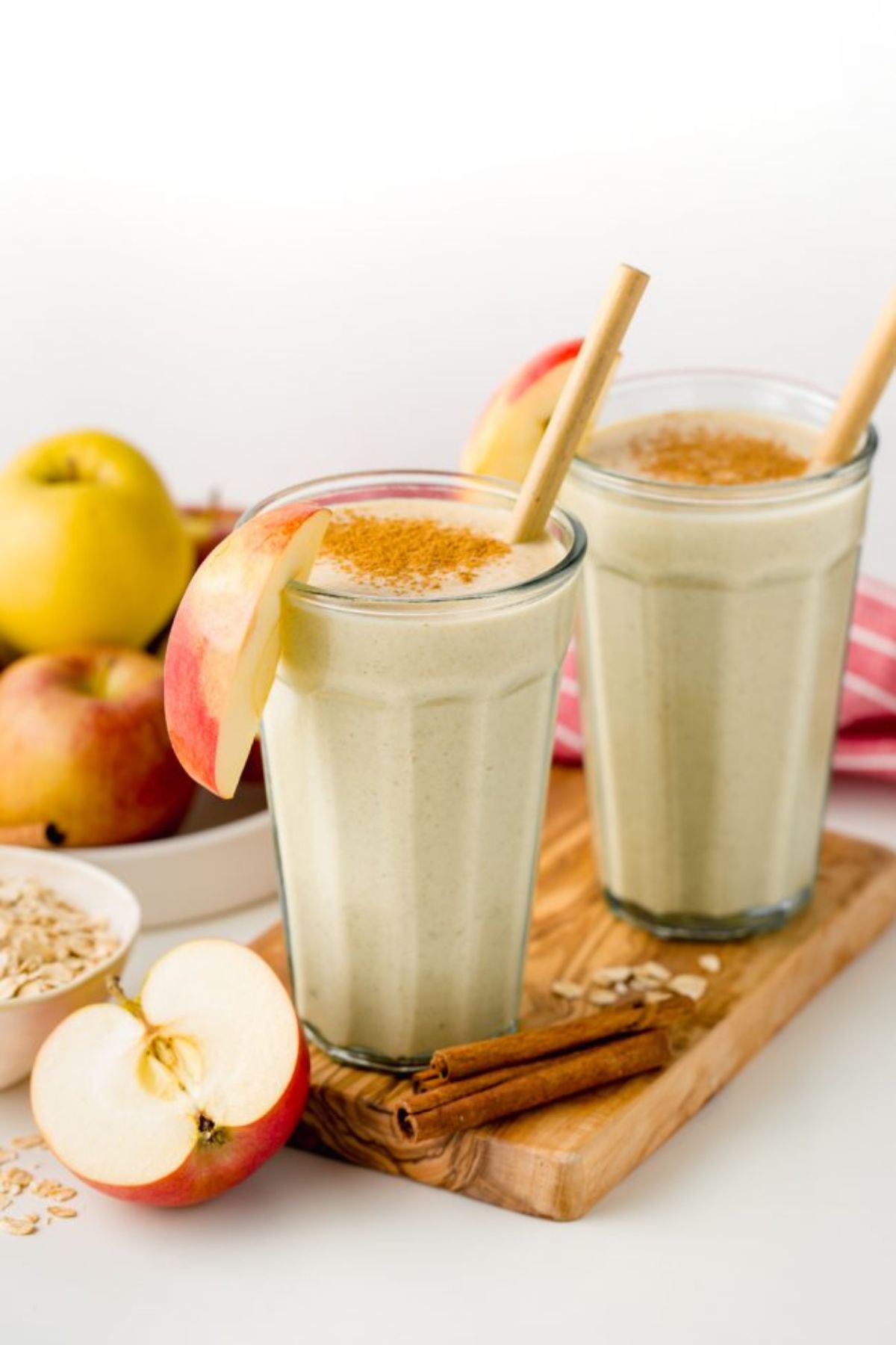 2 glasses of apple pie smoothie on a wooden board, garnished by apple slices, topped with cinnamon and surrounded by cinnamon sticks, apples and oats