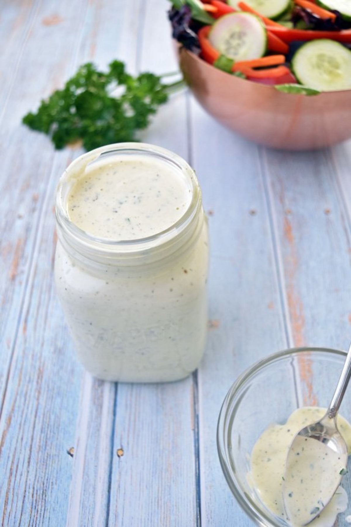 A glass jar of cream dressing on a table next to a bowl of slald, a glass jar with a spoon in it