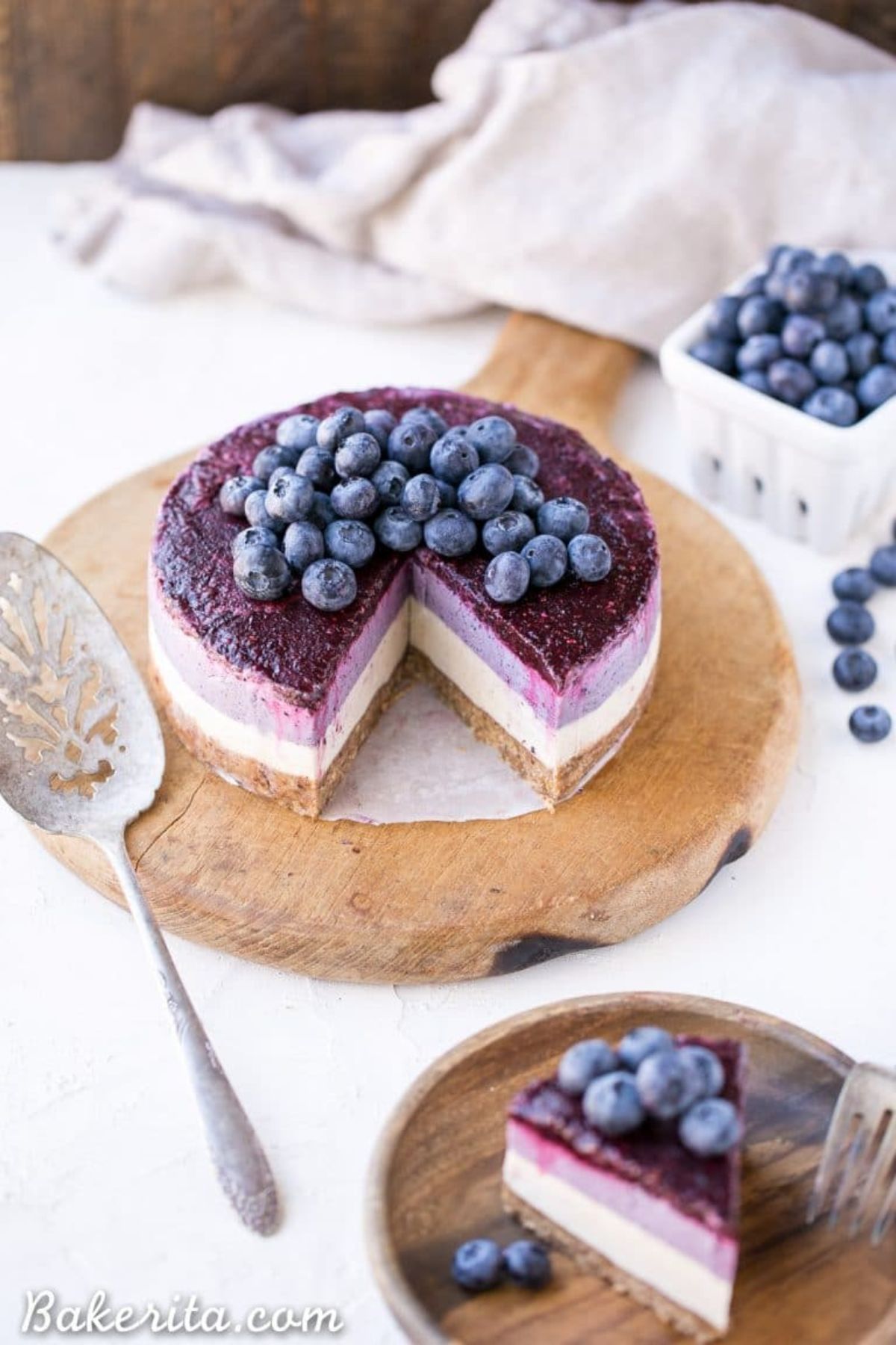 a wooden platter with a layered blueberry cheesecake on it, topped with frsh blueberries. A cake slice is next to it
