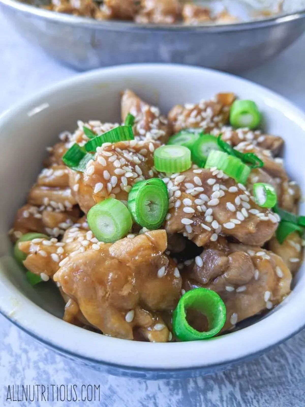 A white bowl with teriyaki chicken sprinkled with sliced scallions and sesame seeds