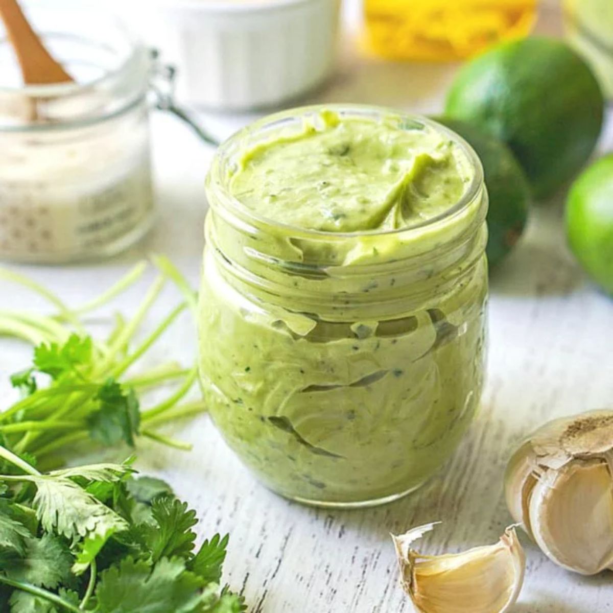 A glass jar ful of thick green dressing. In the foreground is a clove of garlic and some herbs, behind are soem limes and another glass jar