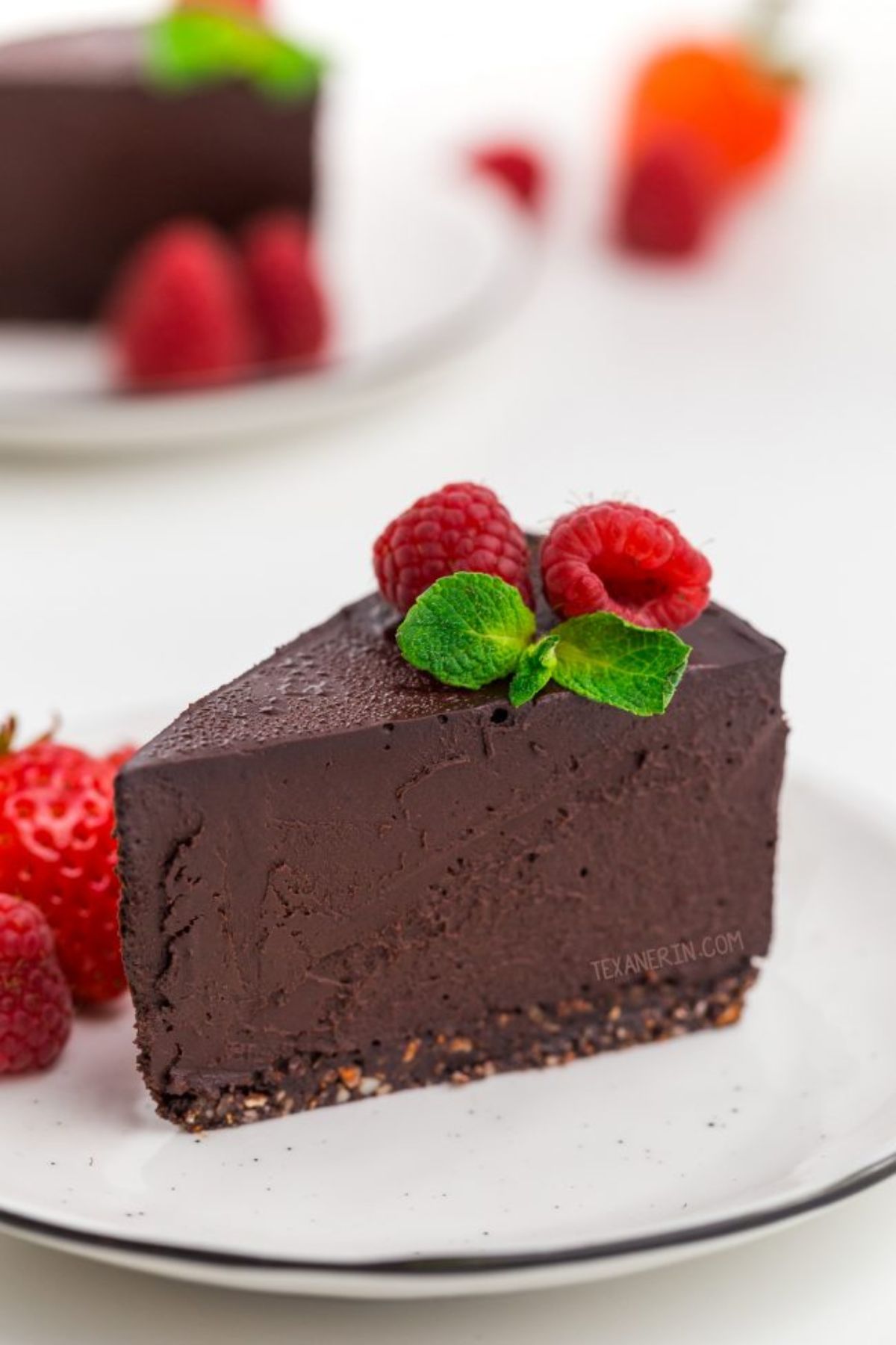 a white plate with a clice of chocolate cheesecake on it, topped with mint leaves and raspberries