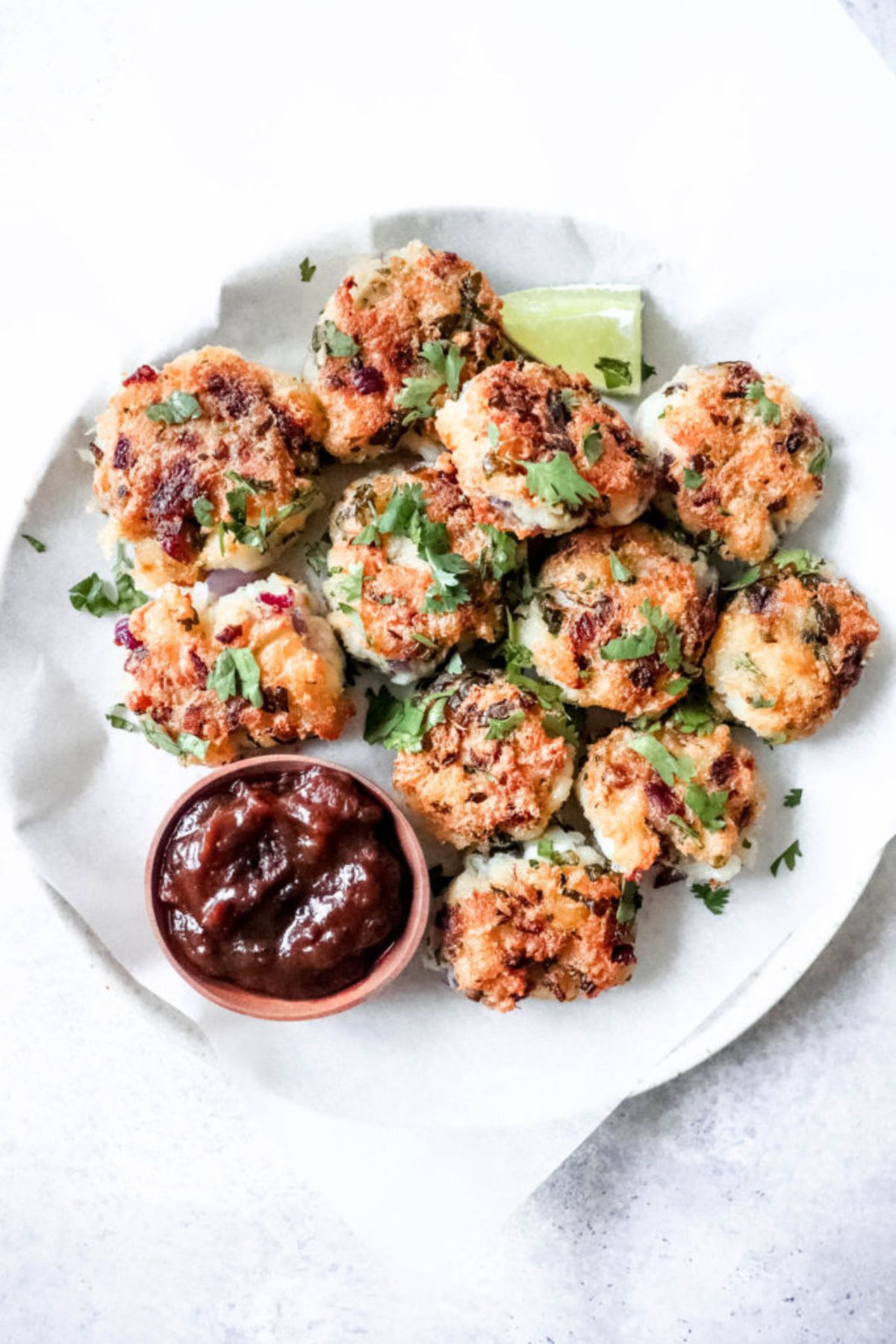 a white plate of cod fritters, sprinkled with chopped herbs, and next to a small bowl of red dipping sauce