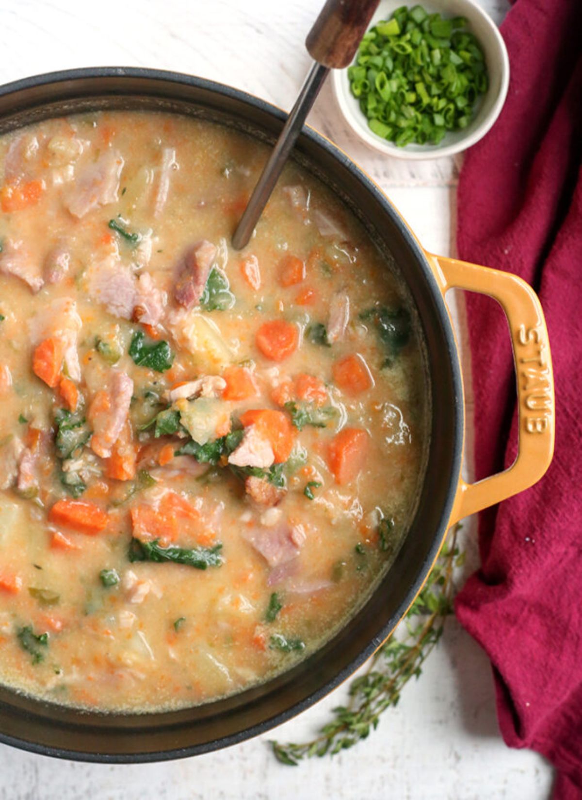 a cast iron casserole dish filled with ham and potato soup with chopped carrots and herbs seen. A spoon is resting in the dish, and a pot of chopped herbs is to the side