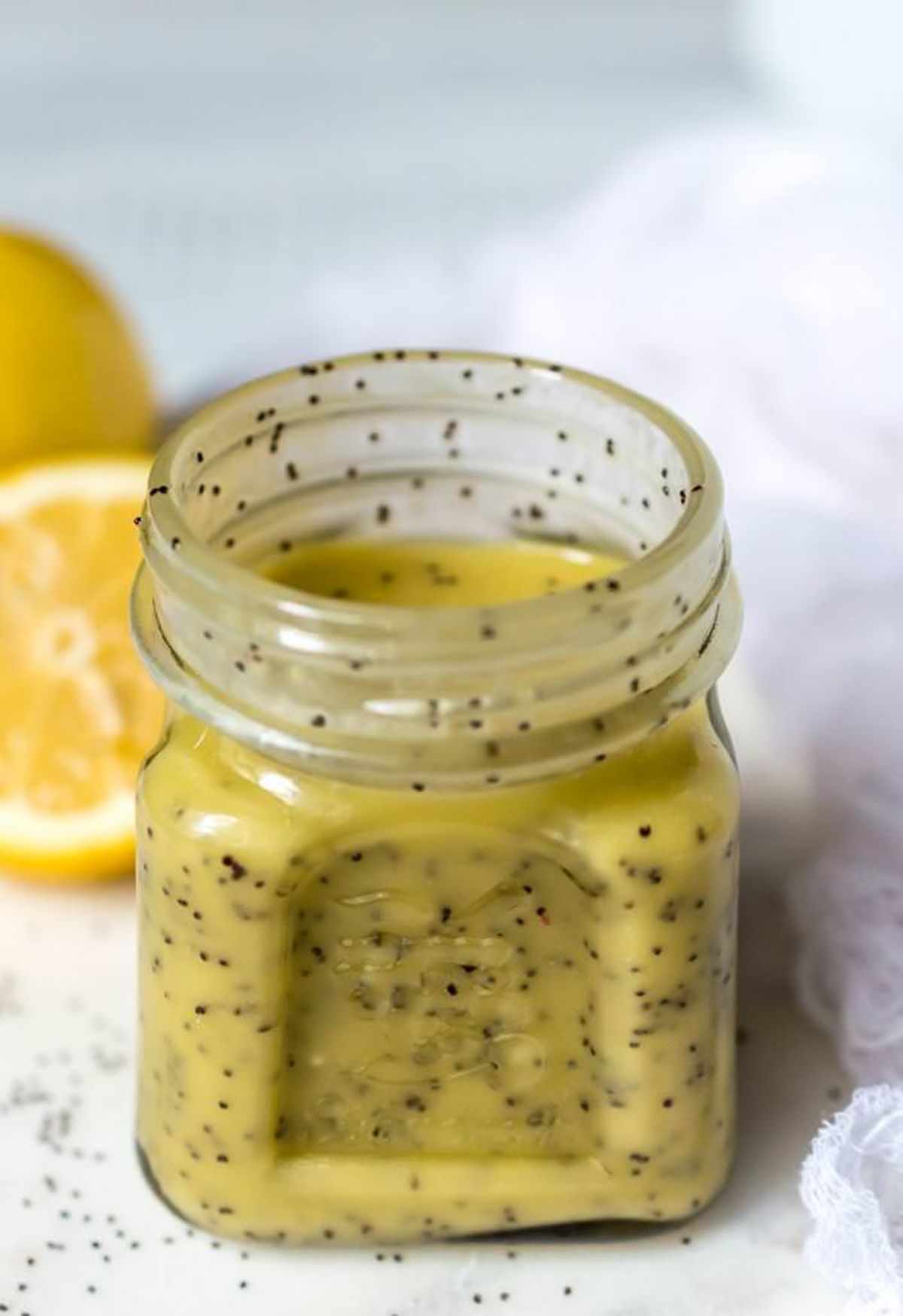 A glass jar of lemon poppy seed dressing with lemon slices behind it