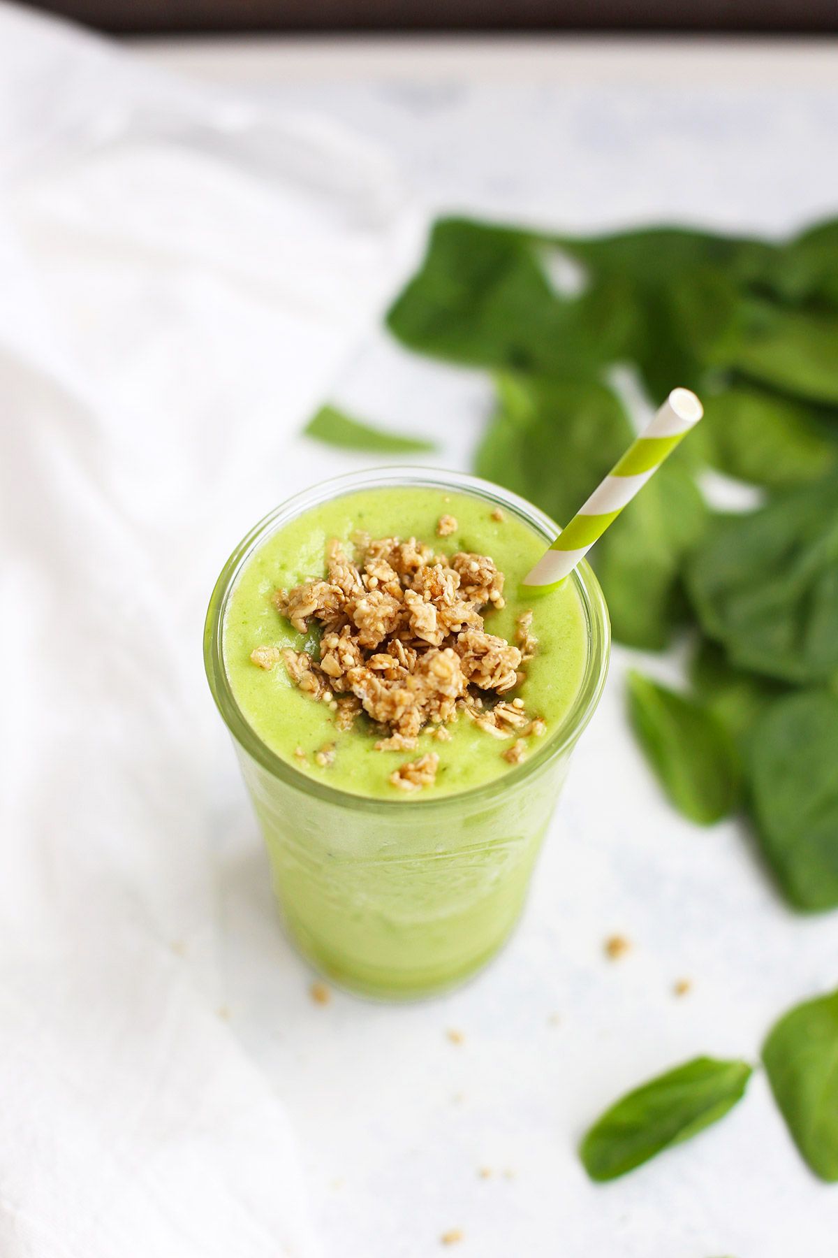 a glass containing pineapple ginger smoothie, topped with crumbled granola and next to a pile of spinach