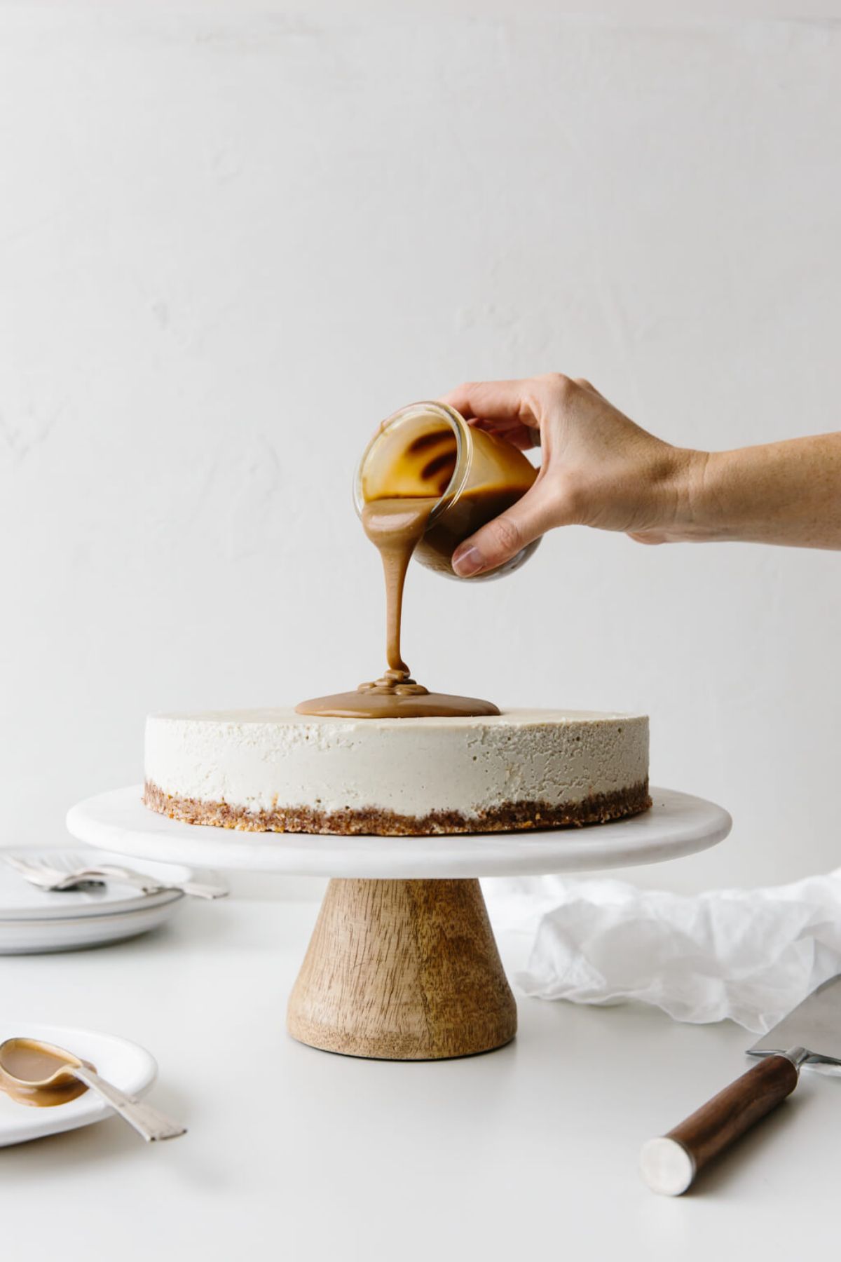 a cake stand with a vanilla cheesecake on it. A hand is pouring caramel sauce from a jar onto the top