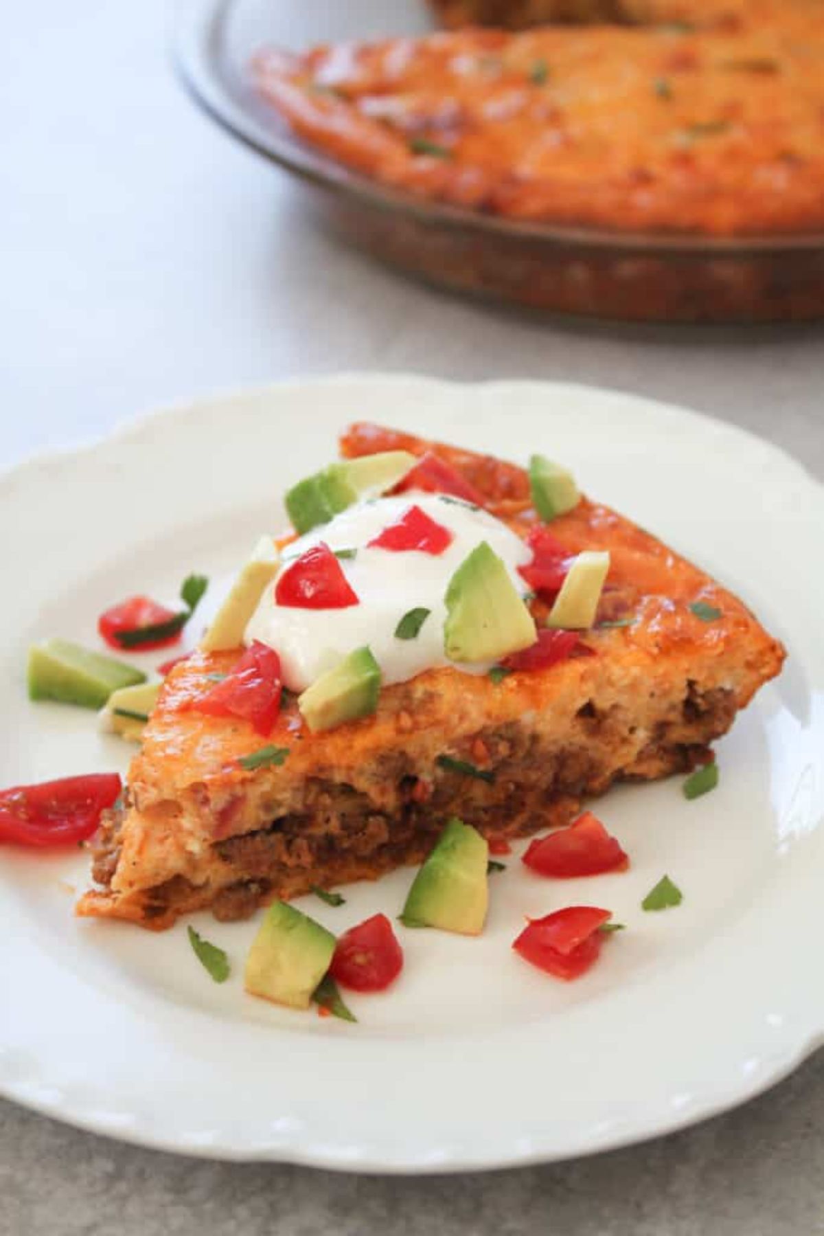 A blurred section of a glass casserole dish with a dark orange contents. In the foreground is a slice of taco pie with avocado, tomato and sour cream topping