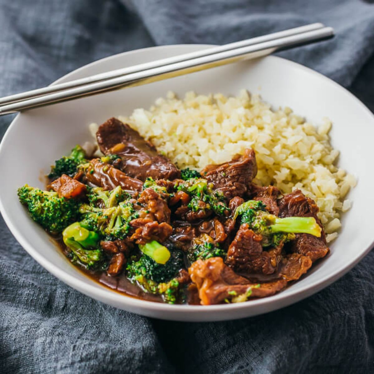 A gray linen cloth lays across the shot. On top is a round white bowl. Inside is a beef and broccoli sauce with cauliflower rice behind it. Silver chopsticks lay across the bowl
