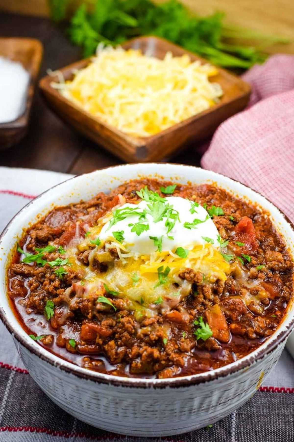 On a dark wooden table is a checked cloth. Blurred in the background is a square wooden bowl full of grated cheese. In the foreground is a round deep bowl filled with chilli, topped with sour cream, cheese and chopped parsley