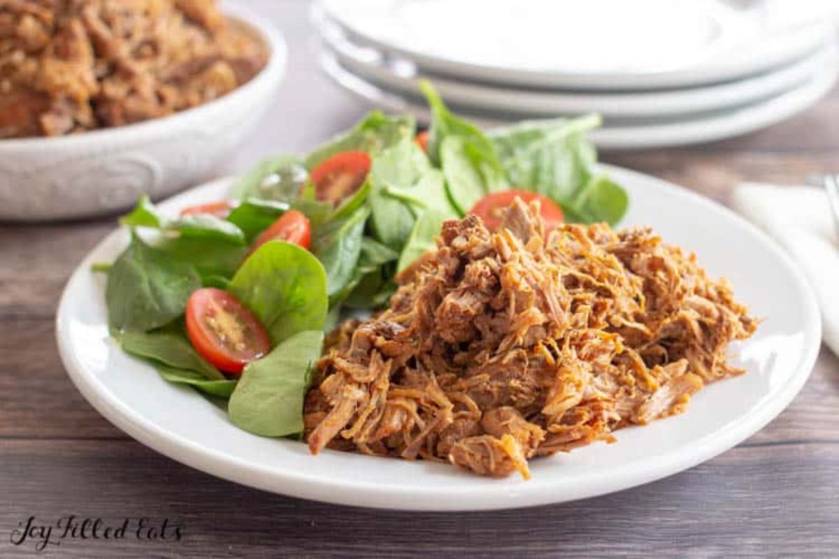 ON a dark wooden table is a partial shot of a deep white bowl filled with a pile of pulled pork, to the right is a pile of white plates. In the foreground is a white plat with half pulled pork and half spinach and tomato salad