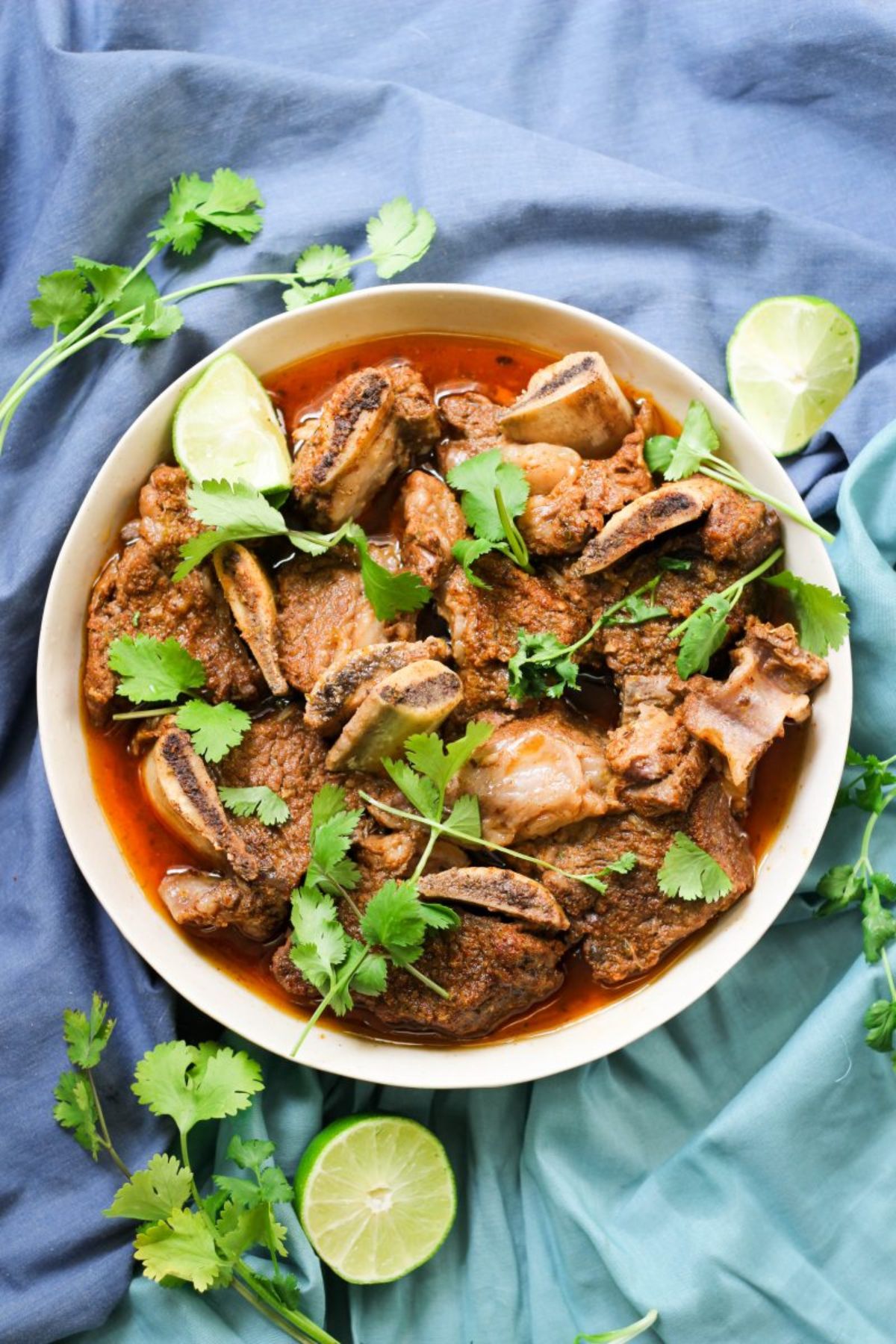 On a blue, ruffled cloth is a round white bowl filled with short rib stew and sprinkled with lime wedges and parsley