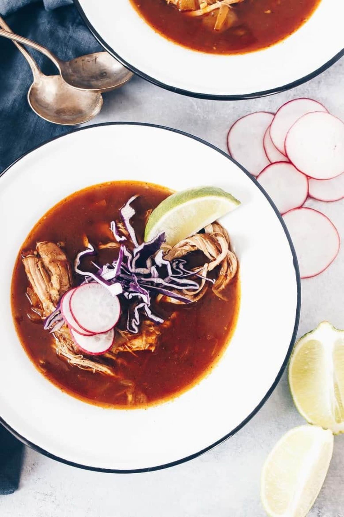 On a white table is sliced radish and lime wedges, with two silver spoons on the top left. A round white soup dish with a blue rim is filled with posole. Topped with sliced radish, sliced red cabbage and a lime wedge
