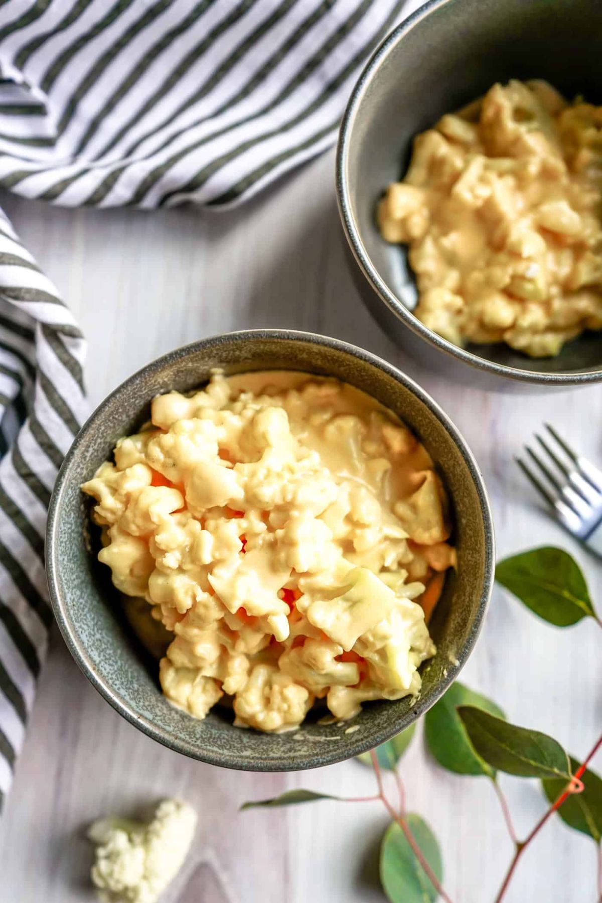 On a white table is a striped rumpled cloth. To the bottom right is some foliage and the top of a silver fork. Two speckled green bowls are full of macaroni cheese.