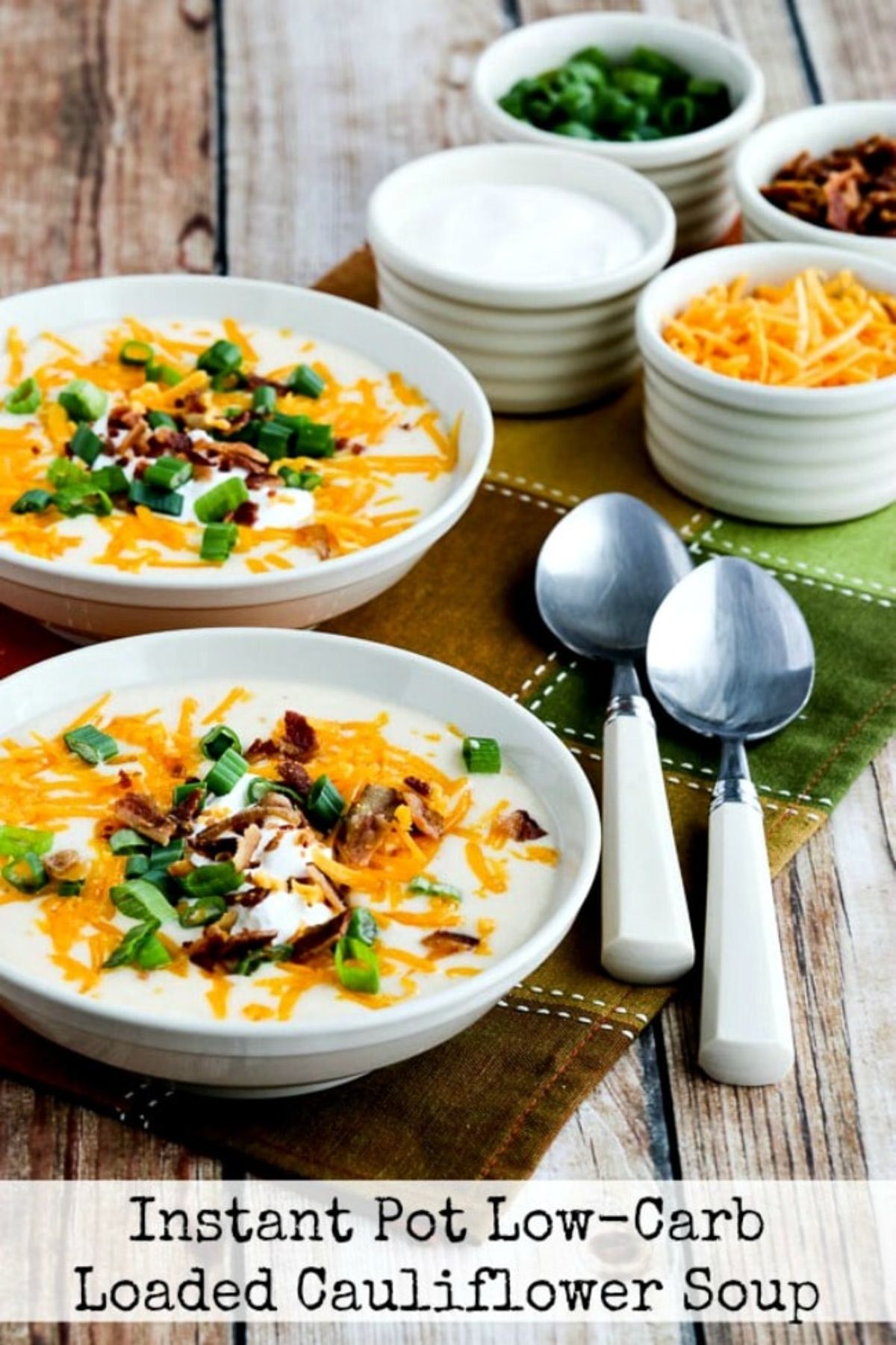 the text reads "Instant Pot Low-Carb Loaded Cauliflower Soup" A wooden table is laden with 4 white small pots filled with grrated cheese, sour cream, chopped scallions, and bacon bits. On a checked napkin are two white handled spoons, and two round white bowls filled with cauliflower soup toppped with cheese, scallions and bacon bits.