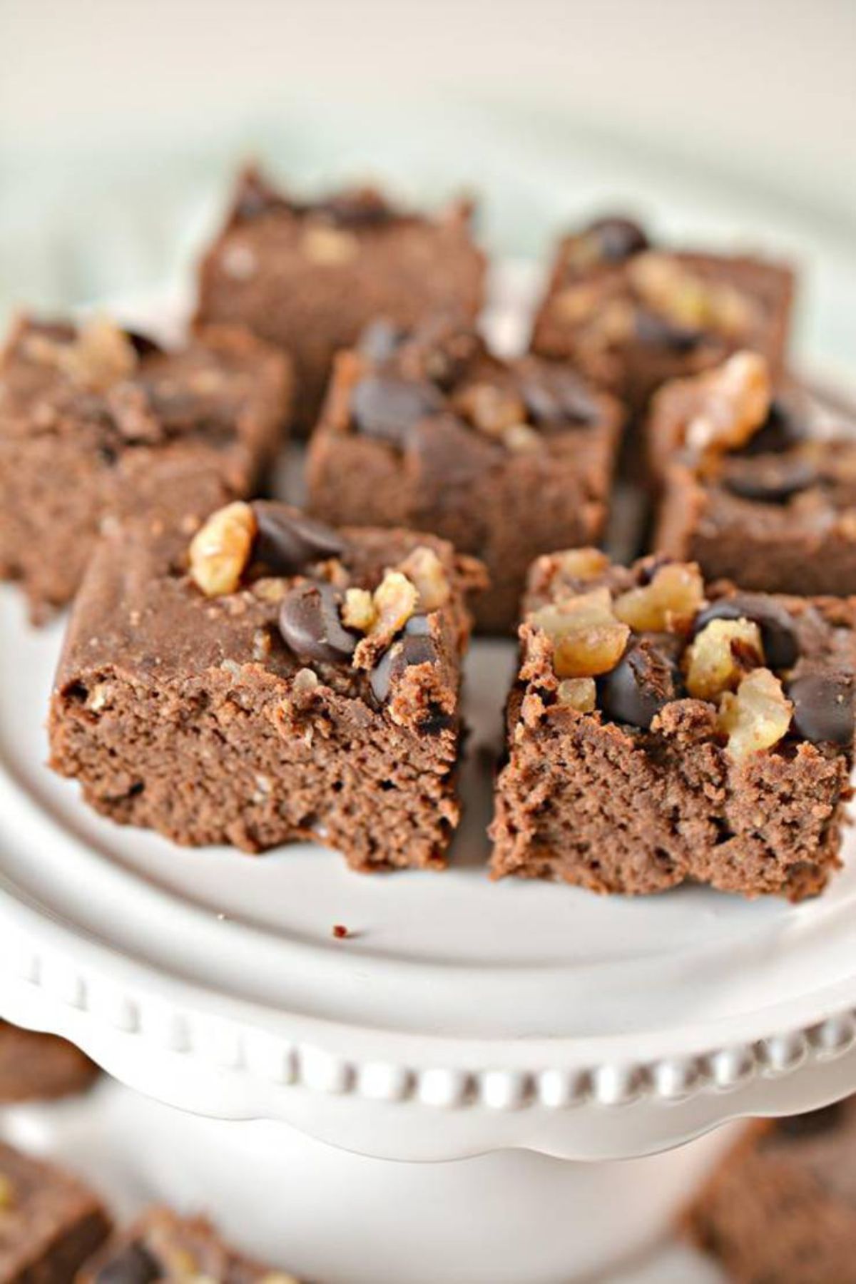 A white porcelain cake stand laden with squares of chocolate brownie topped with chopped nuts and chocolate chips