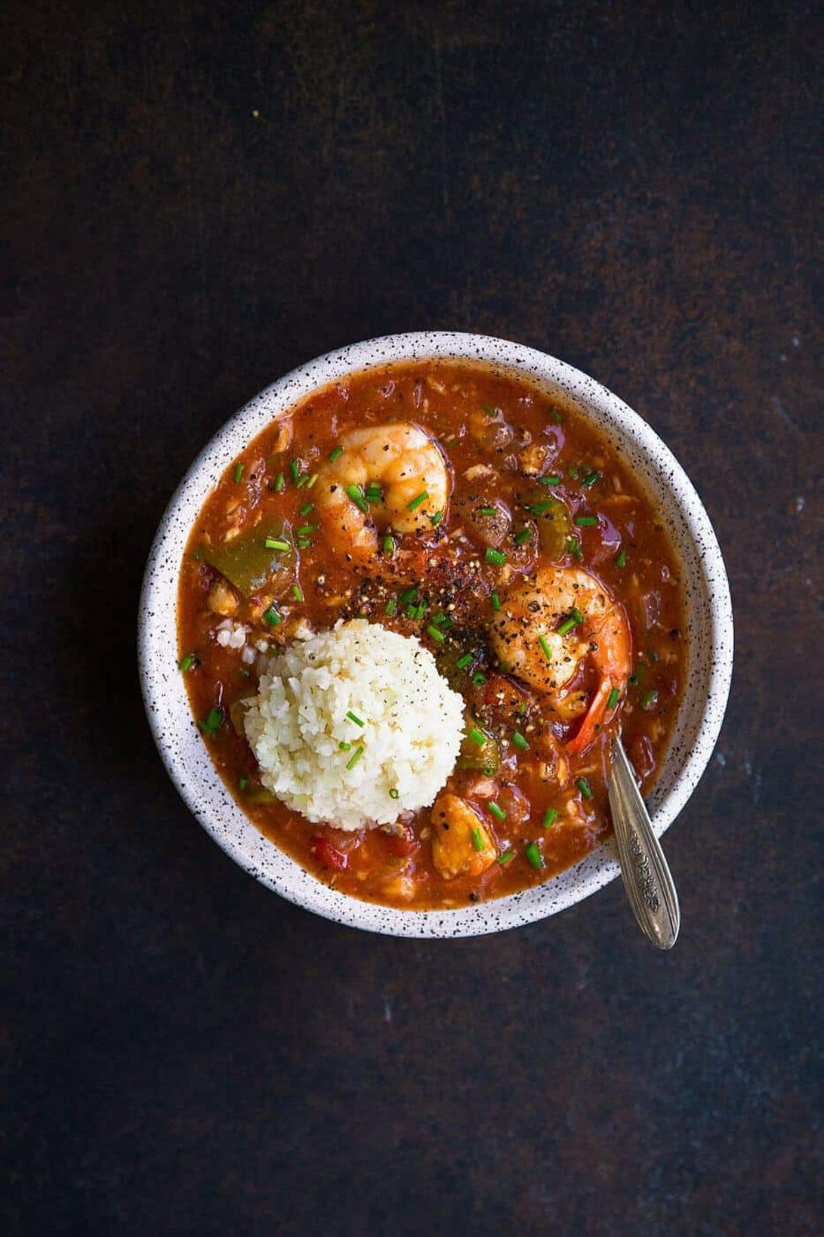 on a dark gray backgrouns is a white bowl filled with shrimp, tomato sauce and cauliflower rice