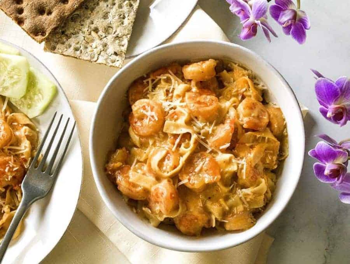 on a white table top is a white bowl filled with cheesy shrimp scampi. Purple flowers are to the right, and to the left is a partial shot of a white plate with the same food and a silver fork on it. Another plate with ryvita crackers is above