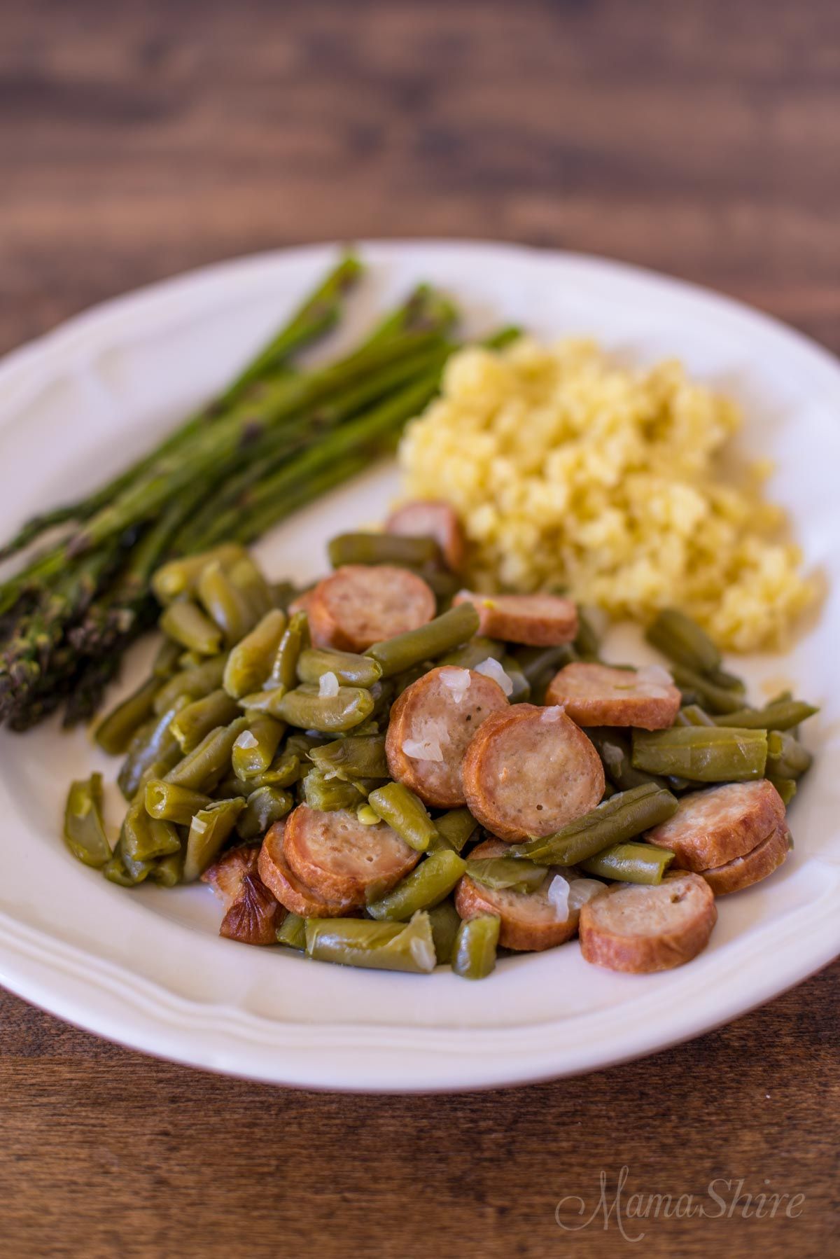 A dark wooden table with a white plate on it. On the plate is a bundle of asparagus, yellow rice, and sausage and beans