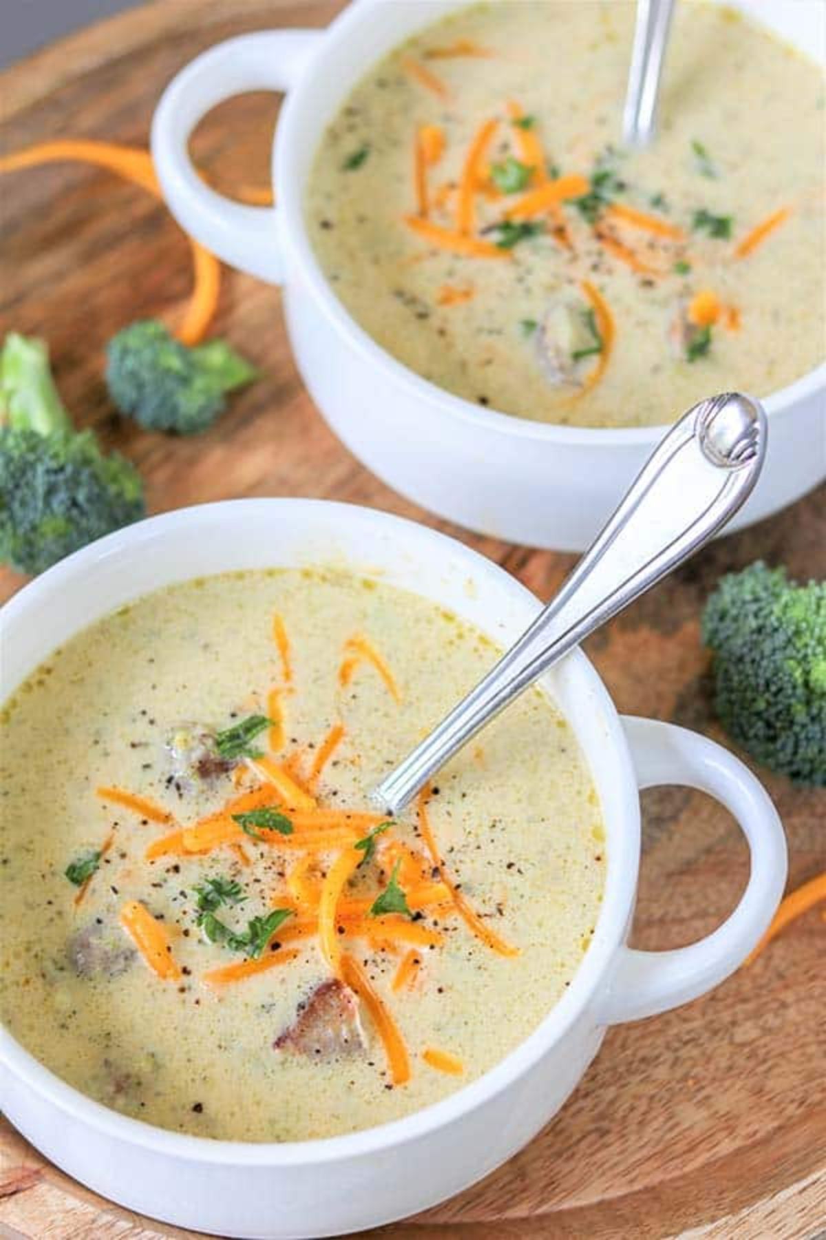 On a wooden board are two round soup bowls with handles. They are filled with a creamy soup, topped with grated orange cheese. Scattered around are small broccoli florets
