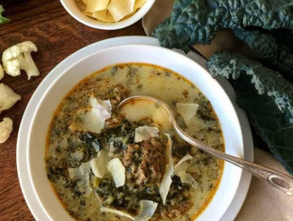 On a wooden table is a white bowl with sausage and kale soup in it, and a silver spoon ain in it. On the right are some leaves of kale, on the left are some florets of cauliflower