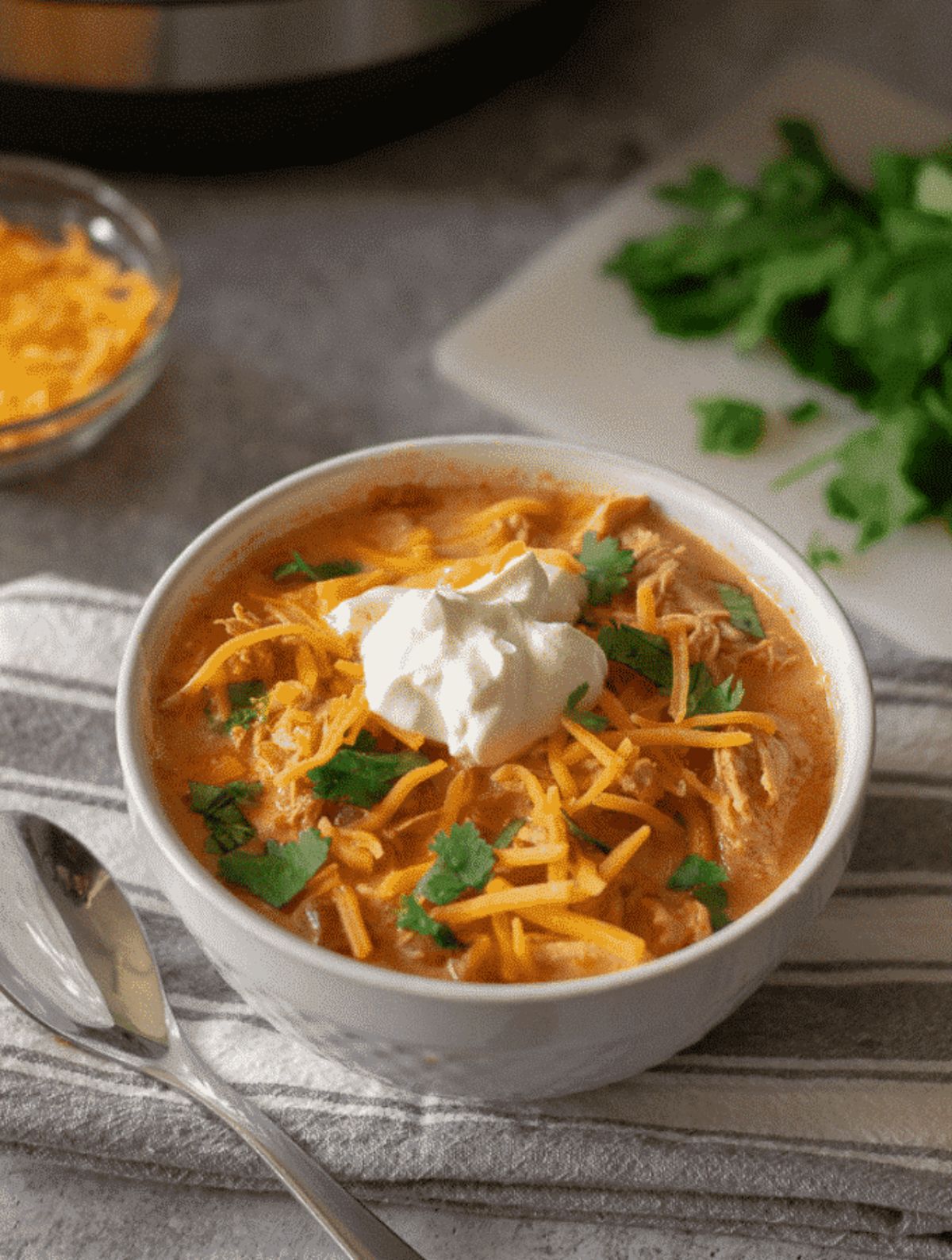 On a gray tabletop, sitting on a striped towel is a white bowl filled with soup, and topped with grated cheese and sour cream