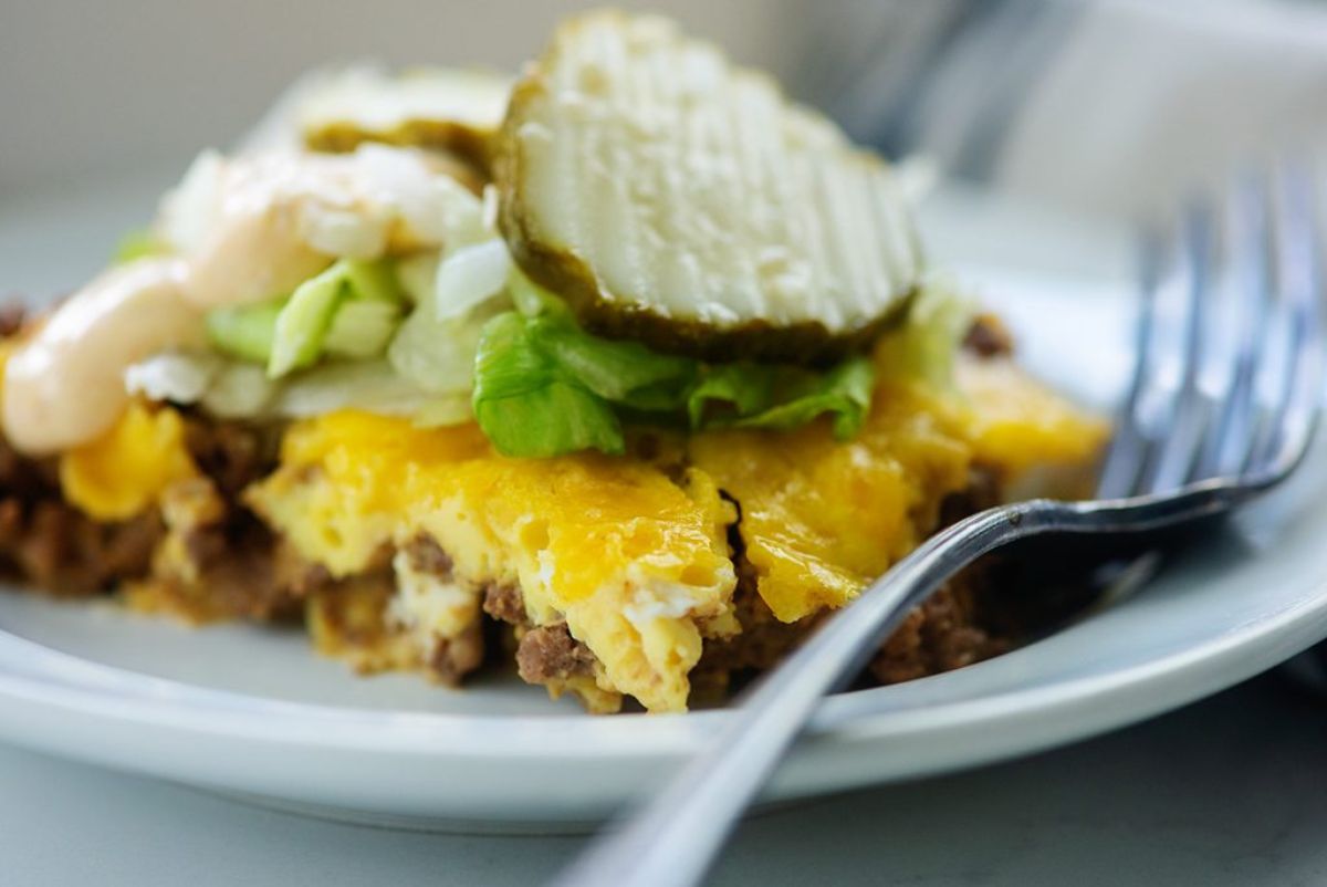 A white plate with a lasagne portion on it, with a pickle garnish. A metal fork sits on the side of the plate