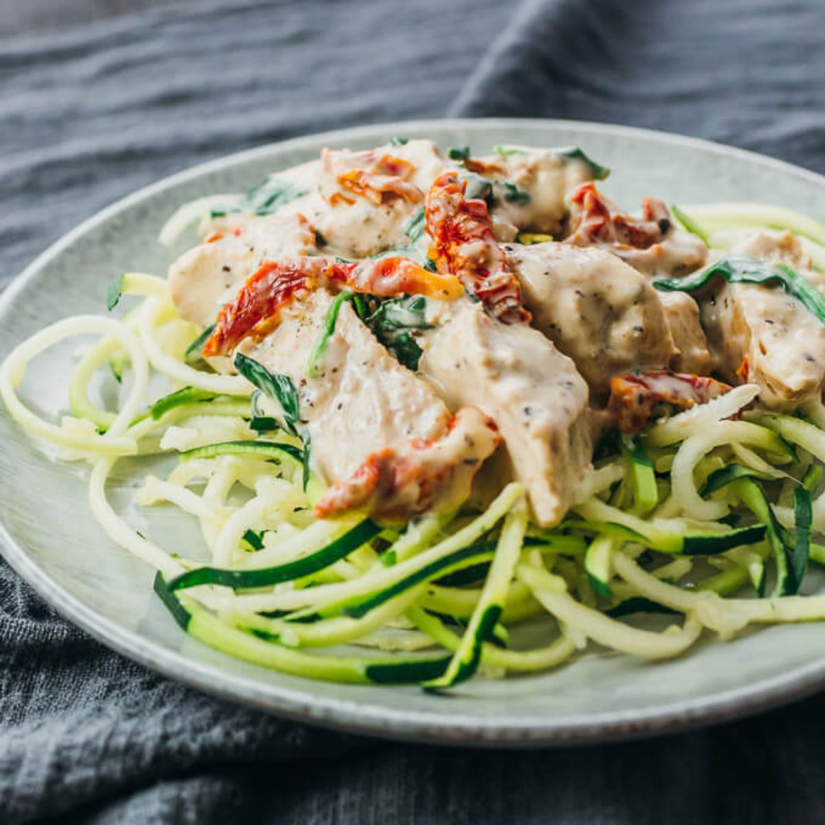 On a dark gray cloth is a white plate. On the plate is a pile of courgetti and then chiken in a creamy sauce with sundried tomatoes