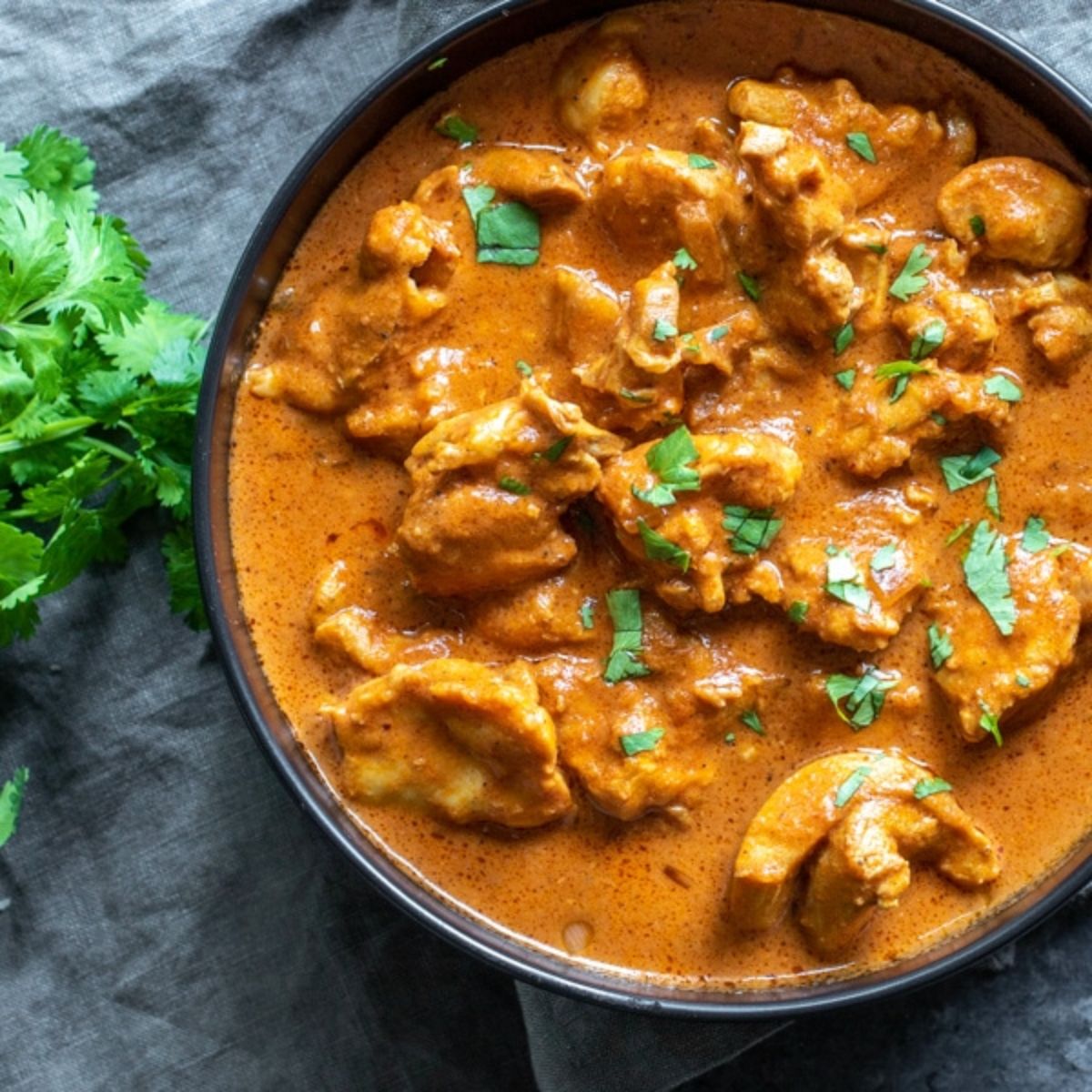 On a light gray cloth next to a pile of coriander is a dark gray bowl filled with butter chicken