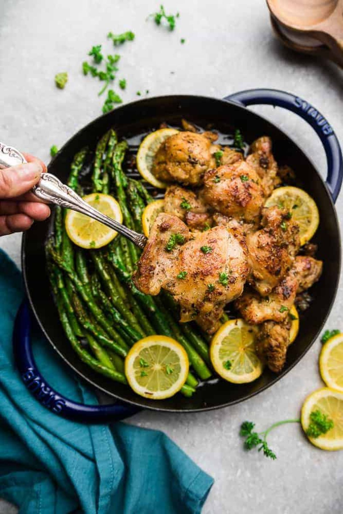 On a light gray surface is a cast iron casserole dish filled with asparagus stalks, chicken and lemon slices. A hand is using a silver spoon to lift out a piece of chicken. Scattered around are chopped herbs and lemon slives