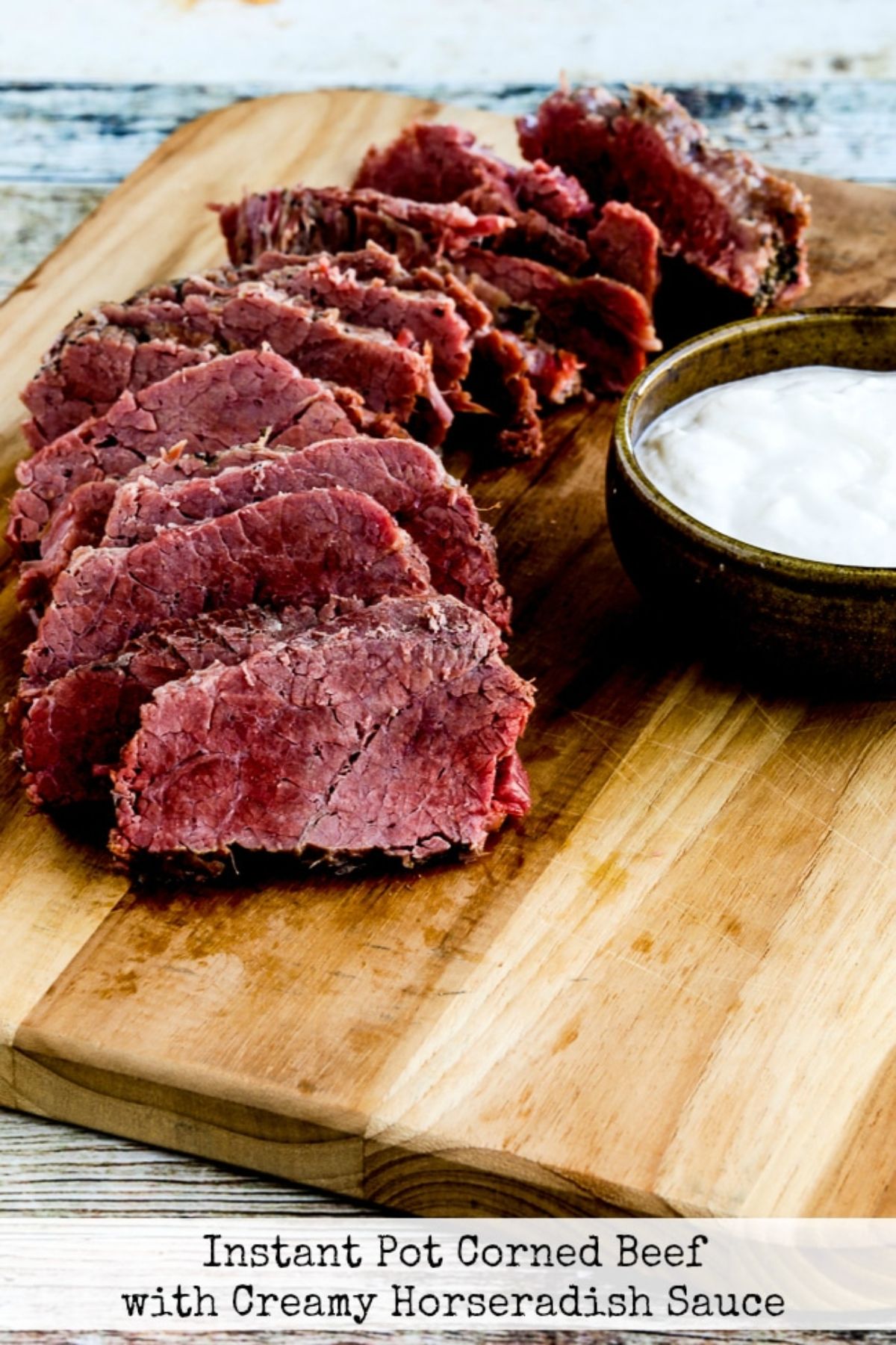 ON a wooden board are slices of beef laid out on top of each other. To the right is a round dish filled with horseradish sauce