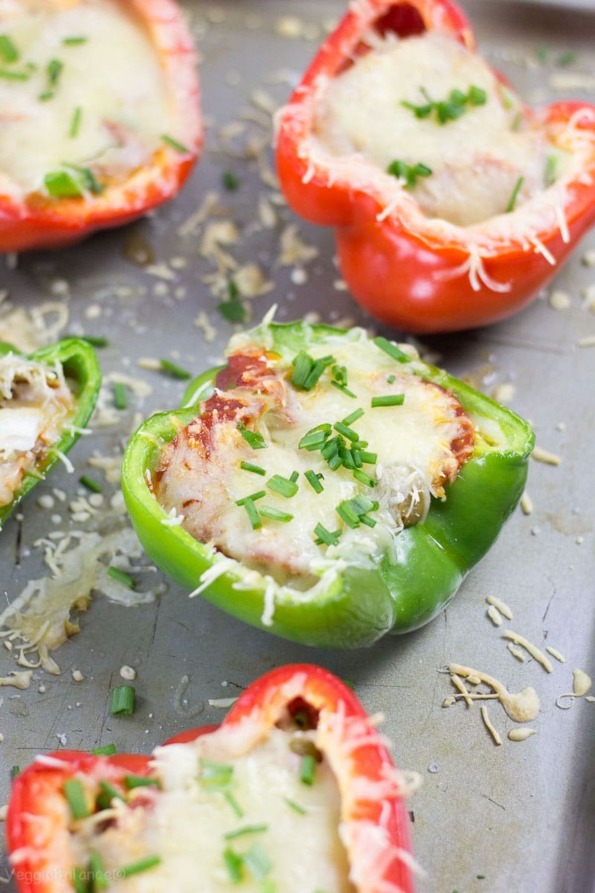 On a gray chopping board are green and red pepper halves filled with cheese mixture and topped with grated cheese and chopped chives