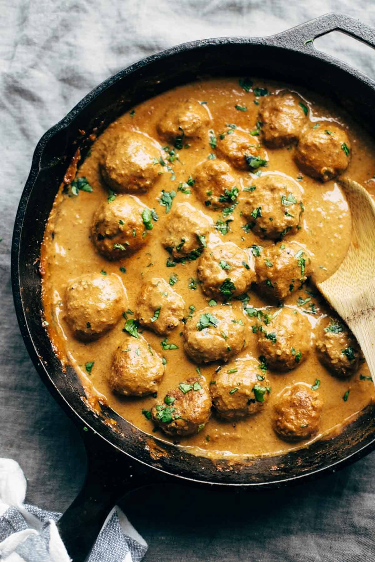 ON a light gray cloth is a cast iron skillet filled with a tomato sauce and meatballs. A wooden spoon is stirring to the right of the pan