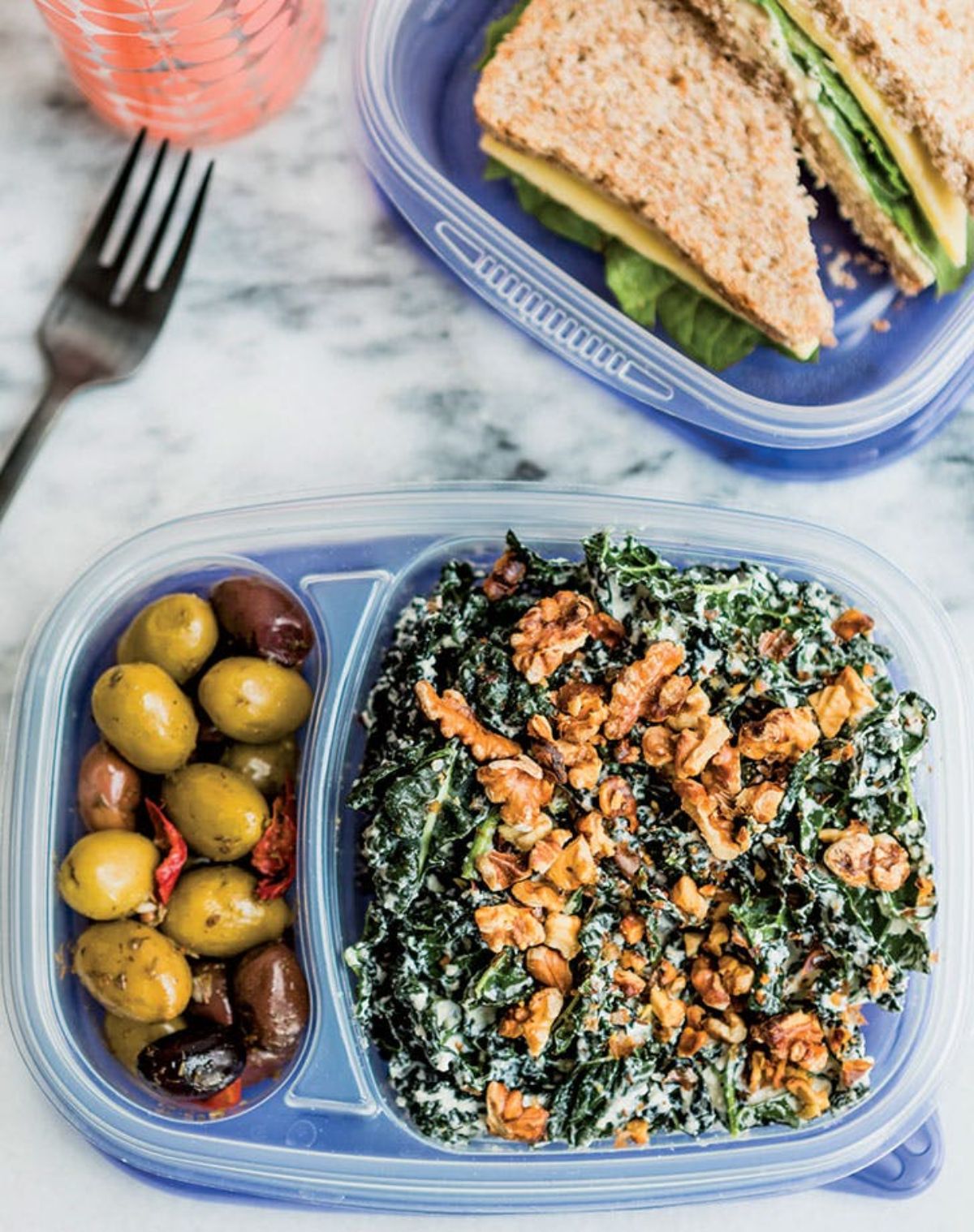 On a white marble countertop are two open blue plastic lunchboxes. Kale salad with walnuts is in the section on the right and mixed olives on the left. The second box behind it is partialy in shot and holds a cheese and lettuce sandwich