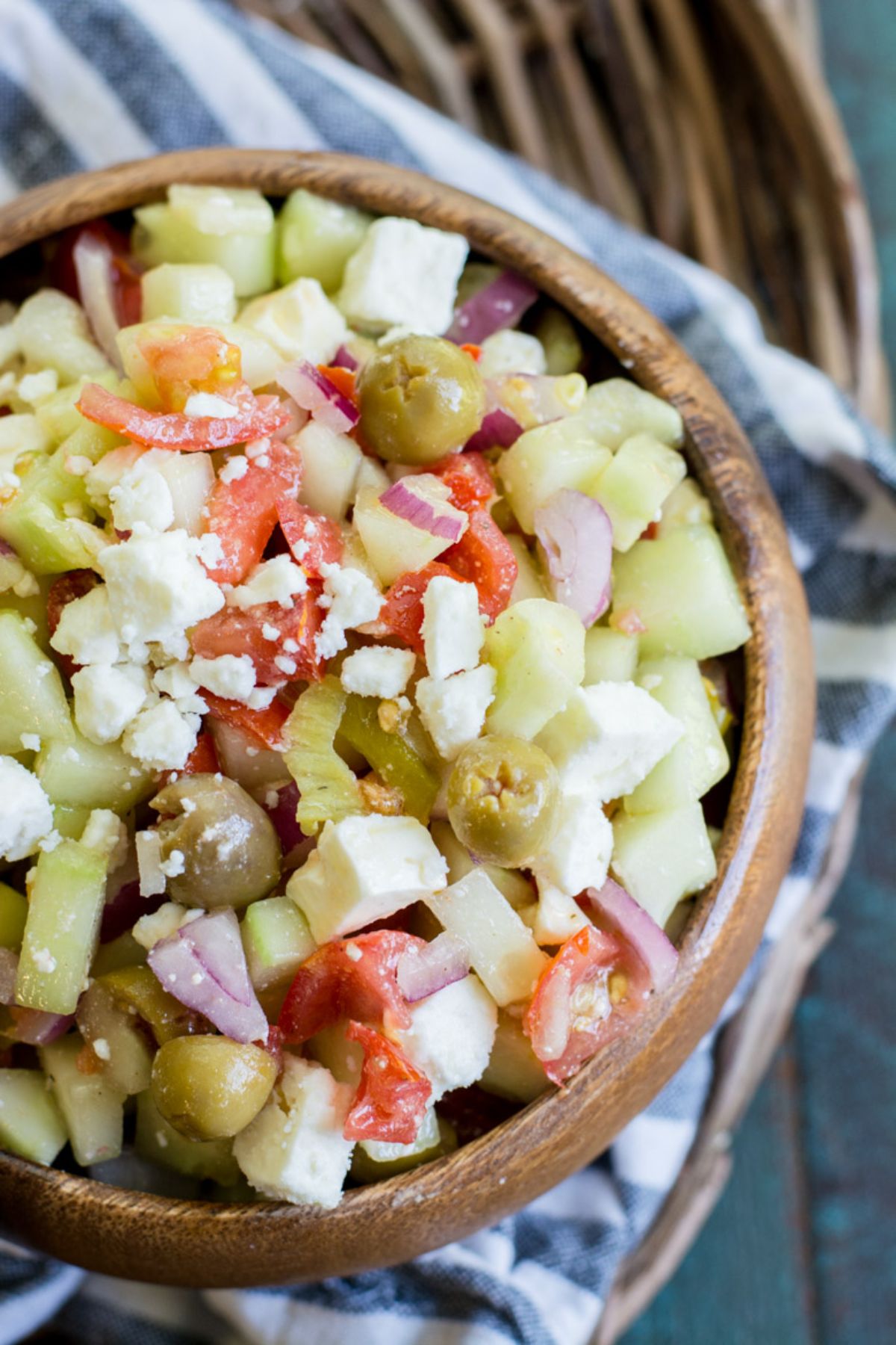 On a blurred wicker table is a bue and white striped cloth. On top of that is a round wooden bowl filled with cucumber salald topped with crumbled feta