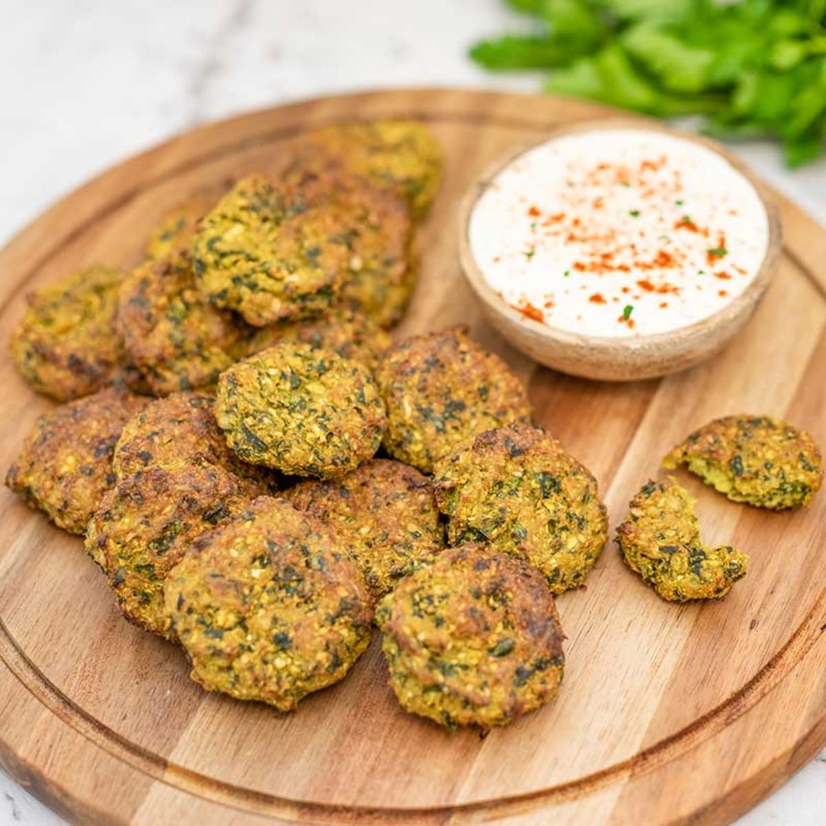 On a whote countertop is a round wooden board. On this is a pile of felafel, and a small bowl of white dip topped with paprika