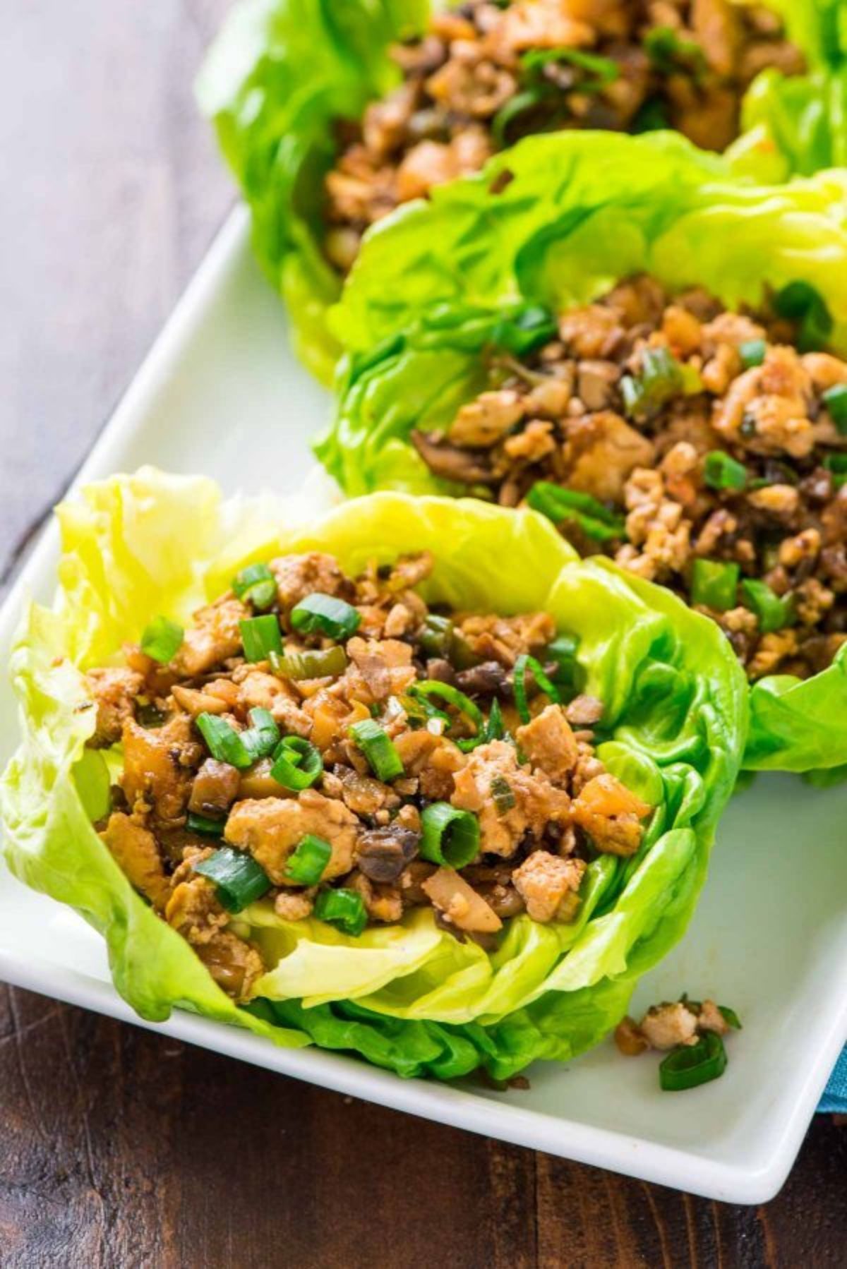 On a dark wooden table is a partial shot of a rectangular white plate. On this are lettuce cups filled with bean and vegetable mix