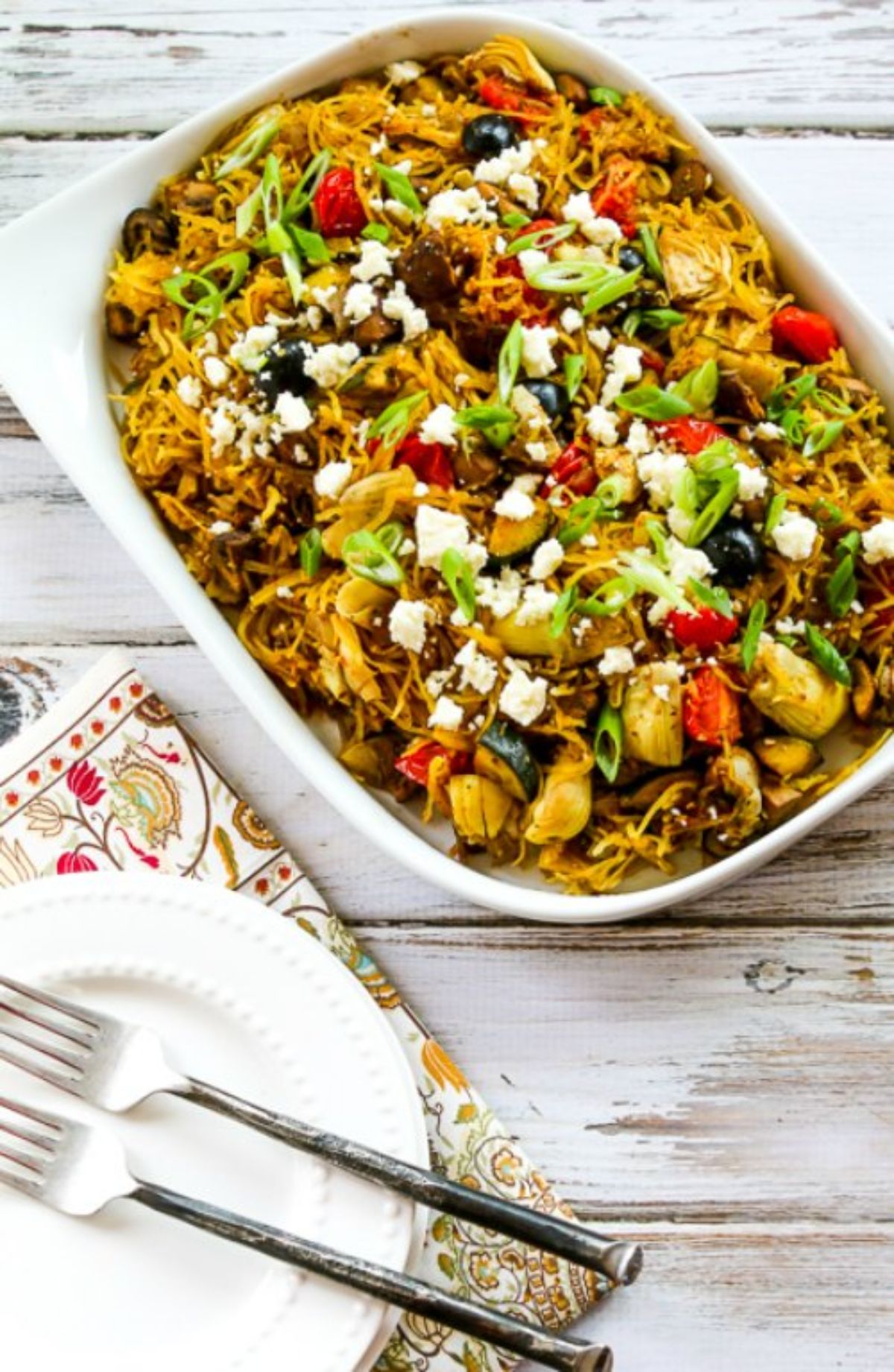 On a whitewashed table is a paisley cloth, with a small white plate and two silver forks on top. Above this is a rectagular white casserole dish filled with spaghetti squash and vegetables